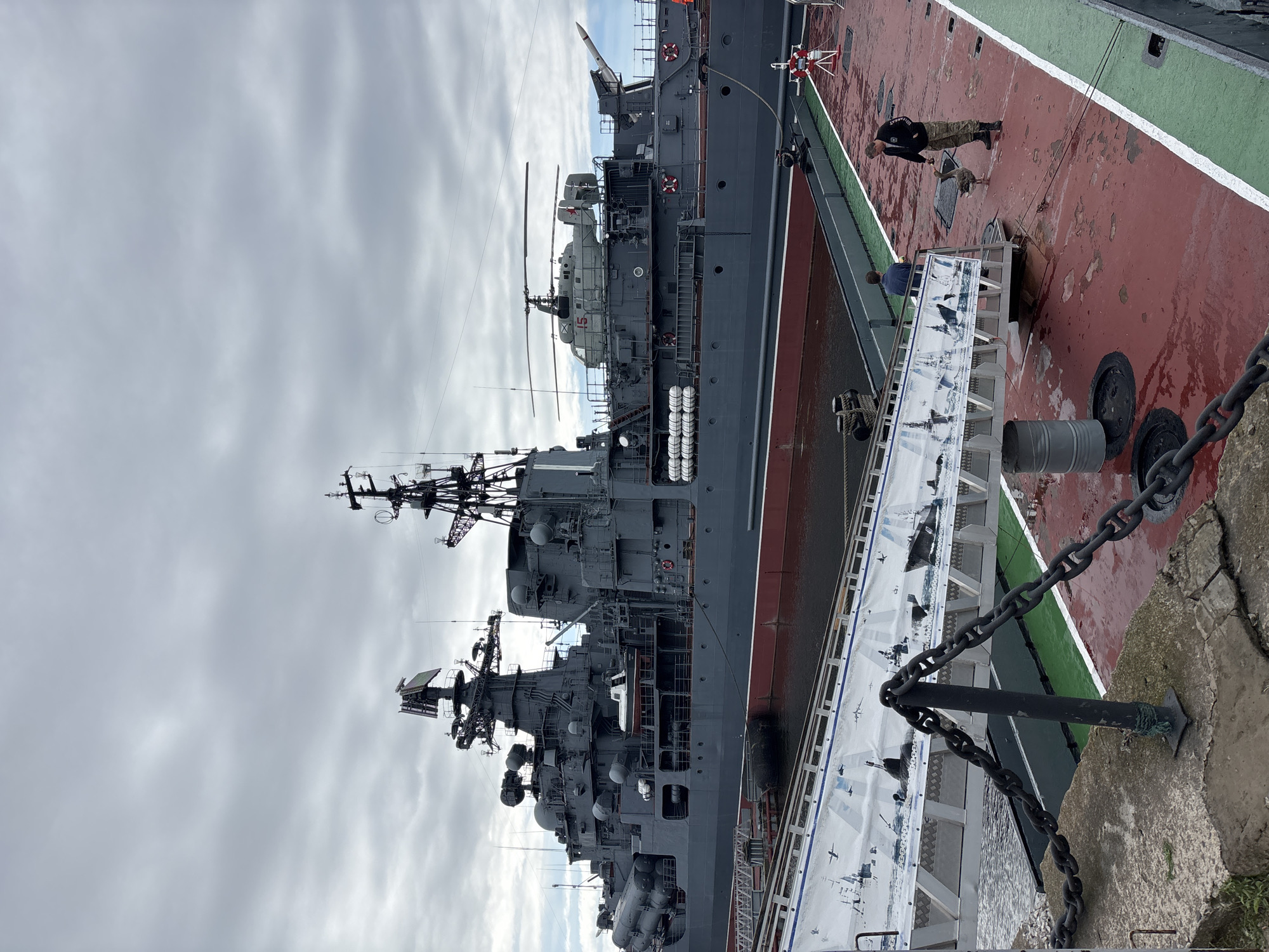 This image, taken in Kronstadt, Russia, depicts a large, dark gray warship docked in a harbor under a cloudy sky. The warship has numerous structures on its deck, including radar equipment, antennas, and what appear to be missile launchers along its side. A helicopter is visible on a platform towards the stern of the ship. In the foreground, a gangway connects the shore to the warship. The gangway is adorned with a banner featuring illustrations of naval vessels and aircraft. To the right of the gangway, on the deck of another vessel, a man in a dark jacket and camouflage pants is interacting with a goose, extending his hand towards it. Another person in a blue shirt stands nearby. The deck is painted red with white and green markings. The scene suggests a naval museum or a working port.