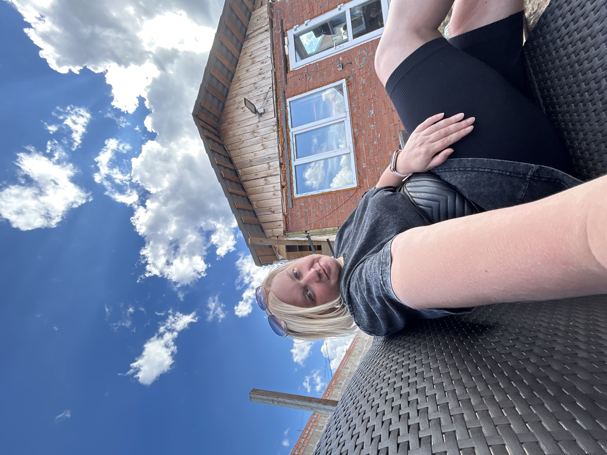 A young woman with blonde hair and sunglasses resting on her head is captured in a selfie. She is wearing a dark grey t-shirt and black biker shorts. Her right hand is placed on her thigh, and she is leaning on a dark-colored, woven-textured surface, possibly outdoor furniture.
In the background, a partially constructed brick building is visible, with wooden planks extending from the roofline and windows reflecting the sky. The building appears to be in a state of disrepair or ongoing construction. The sky is a vibrant blue, dotted with fluffy white clouds, suggesting a sunny day. The image is oriented sideways, with the woman appearing to be lying down or sitting and looking up.
The scene suggests an outdoor setting, possibly a backyard or construction site, during daytime given the bright sunlight and clear sky. The woman's expression is neutral. No visible text is present in the image. The context provided places this scene in Chaikovsky, Russia.