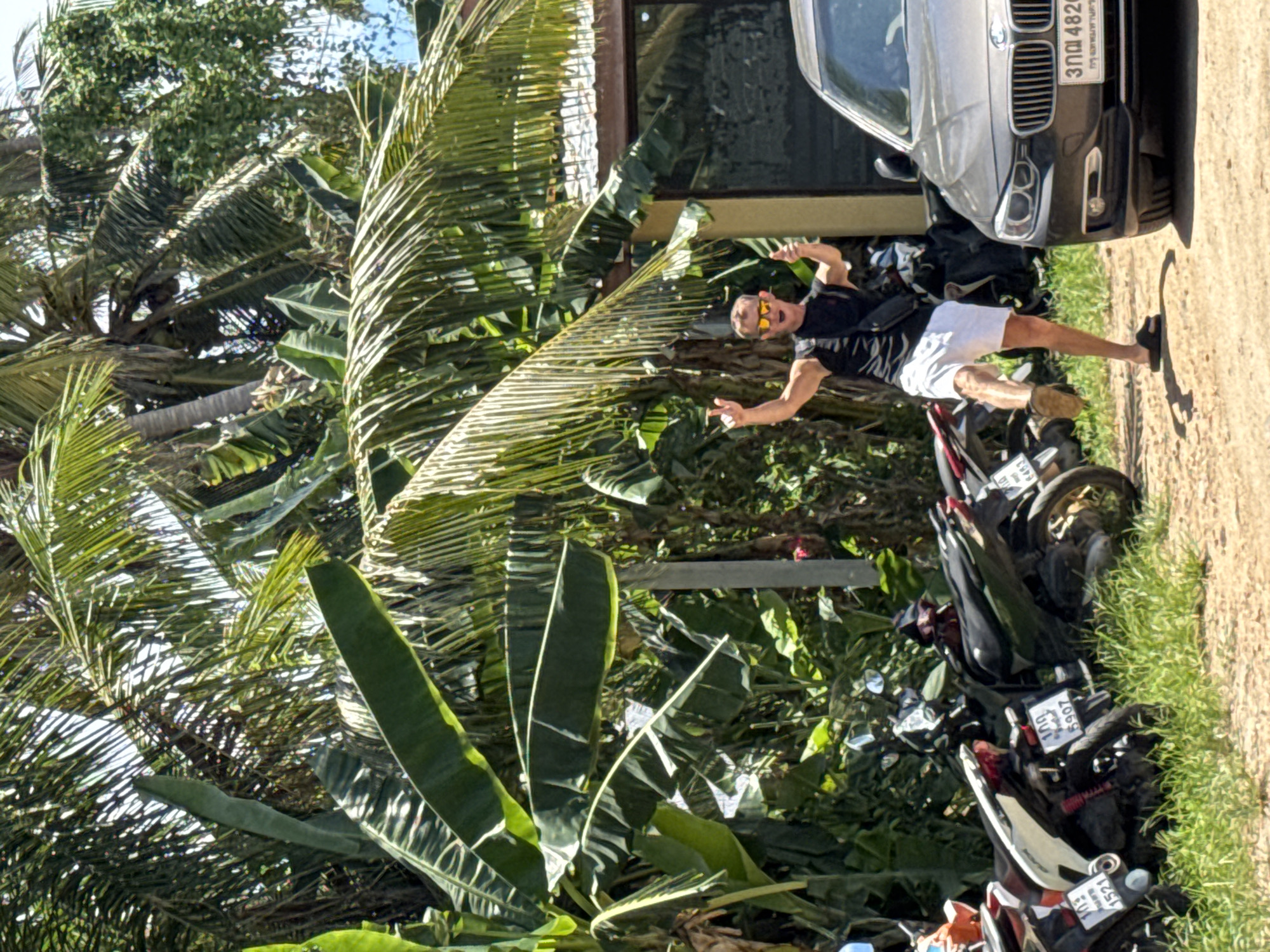 In ตำบลเกาะพะงัน, Thailand, a man in a black t-shirt and white shorts is standing with his arms raised in a celebratory pose. He is wearing sunglasses and sandals, and a black fanny pack is slung across his chest. He is in front of a row of parked motorcycles and a dark-colored BMW sedan. The background features lush green palm trees and other tropical foliage. The ground is a mixture of dirt and grass. The man's posture suggests he is experiencing joy or excitement.