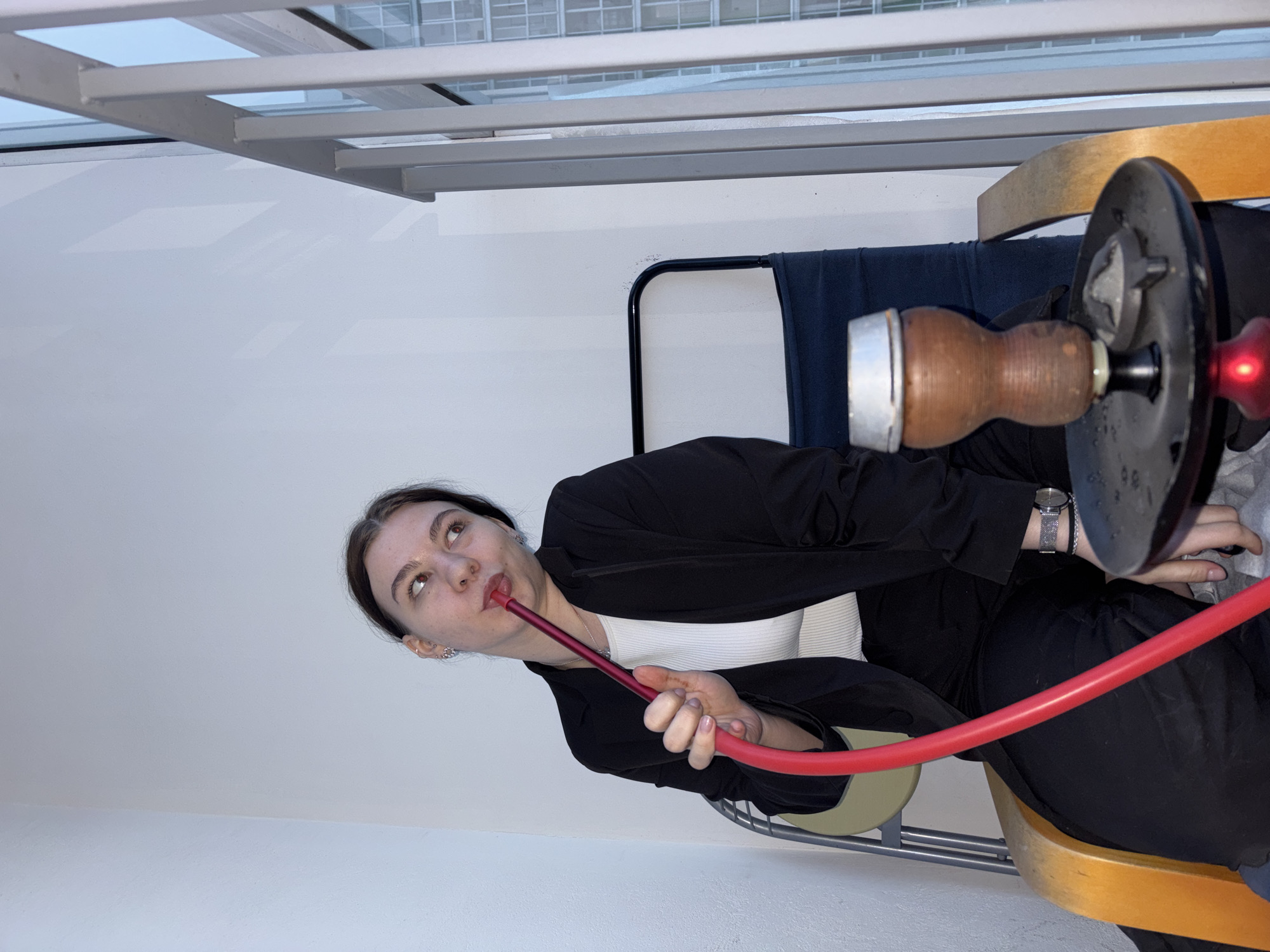 A woman is seated in a chair, smoking from a hookah. She is wearing a white top and a black blazer. She has dark hair pulled back from her face. A watch is visible on her left wrist. The hookah has a red hose and a wooden mouthpiece. The chair has a wooden seat and a metal frame. A window is visible in the background. The location could be Perm, Russia.