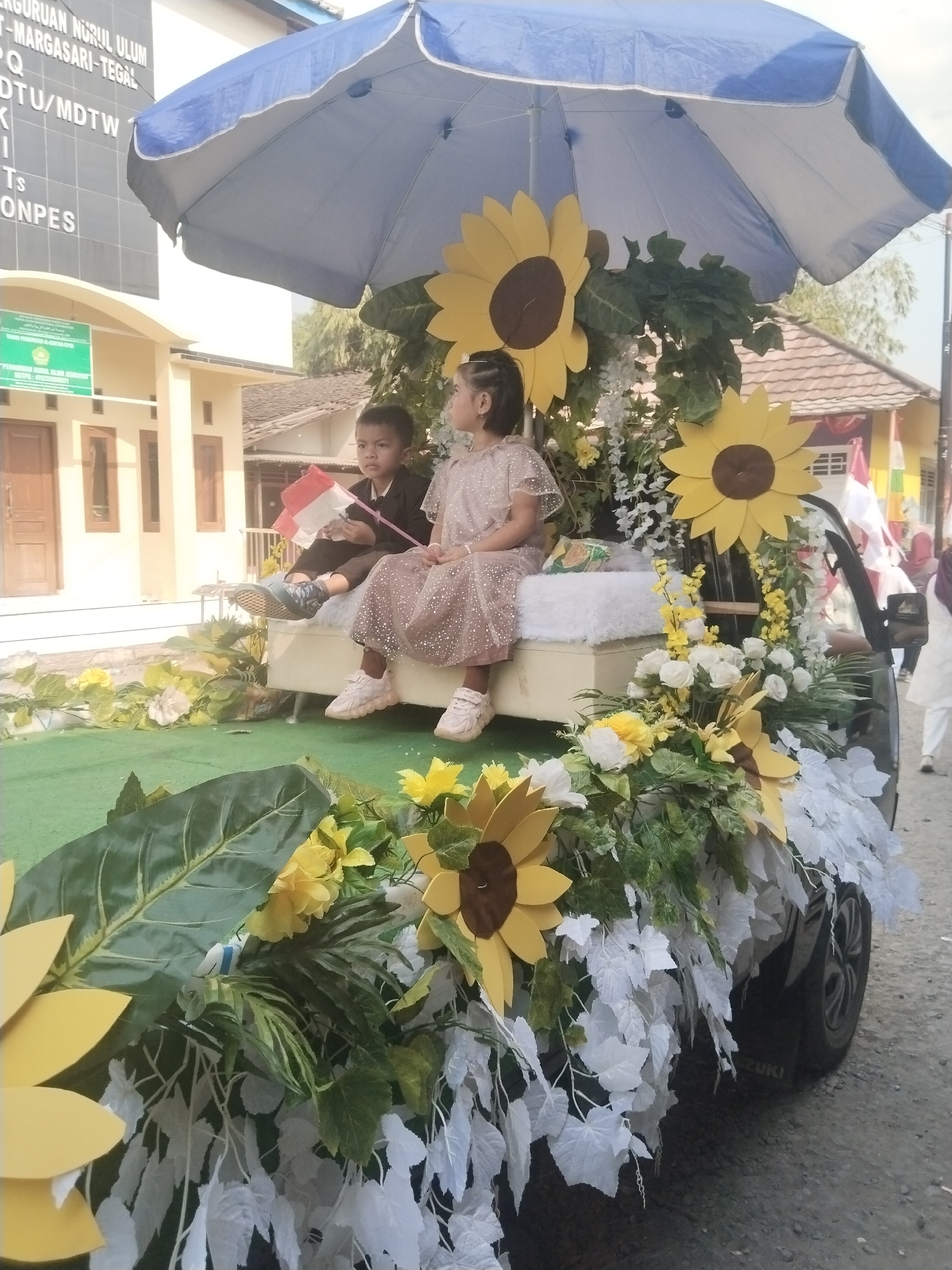 The image depicts a decorated float, likely part of a parade in Jembayat, Indonesia. Two young children, a boy and a girl, are seated on a white cushioned platform under a large blue and white umbrella. The boy, dressed in a dark jacket and pants, holds a small Indonesian flag. The girl wears a light-colored, sparkly dress. The float is adorned with artificial sunflowers, green foliage, and white artificial flowers. In the background, buildings with tan walls and brown roofs are visible. The street is paved with asphalt, and a portion of the float's wheel and the branding "SUZUKI" on the vehicle can be seen. The overall scene suggests a festive and celebratory event.