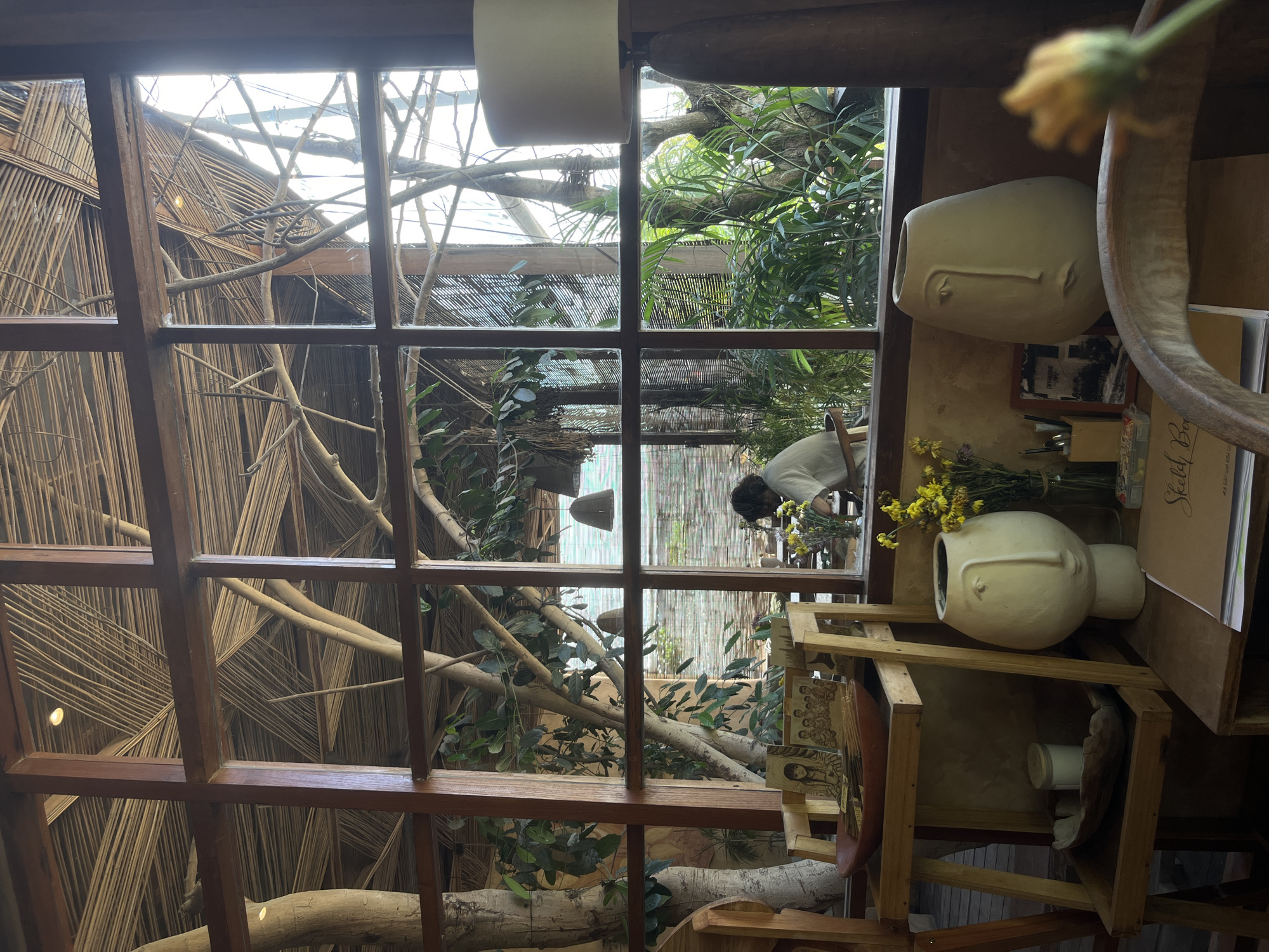The image depicts an indoor scene in what appears to be a building with a rustic, natural aesthetic. The view is framed by a multi-paned window with a dark wooden frame.
Outside the window, there's a structure or enclosure featuring thatched or woven bamboo panels, along with trees, branches, and various plants, creating a garden-like environment. A person wearing a light-colored shirt is partially visible outside, tending to the plants.
Inside the building, near the window, is a wooden shelf or stand holding various objects. These include two white, face-shaped vases, one with yellow flowers in it. There are also books, papers, and other decorative items arranged on the shelf.
The overall style suggests a bohemian or nature-inspired cafe or studio space. There is no indication that this location is in Kuta Selatan, Indonesia, despite the request.