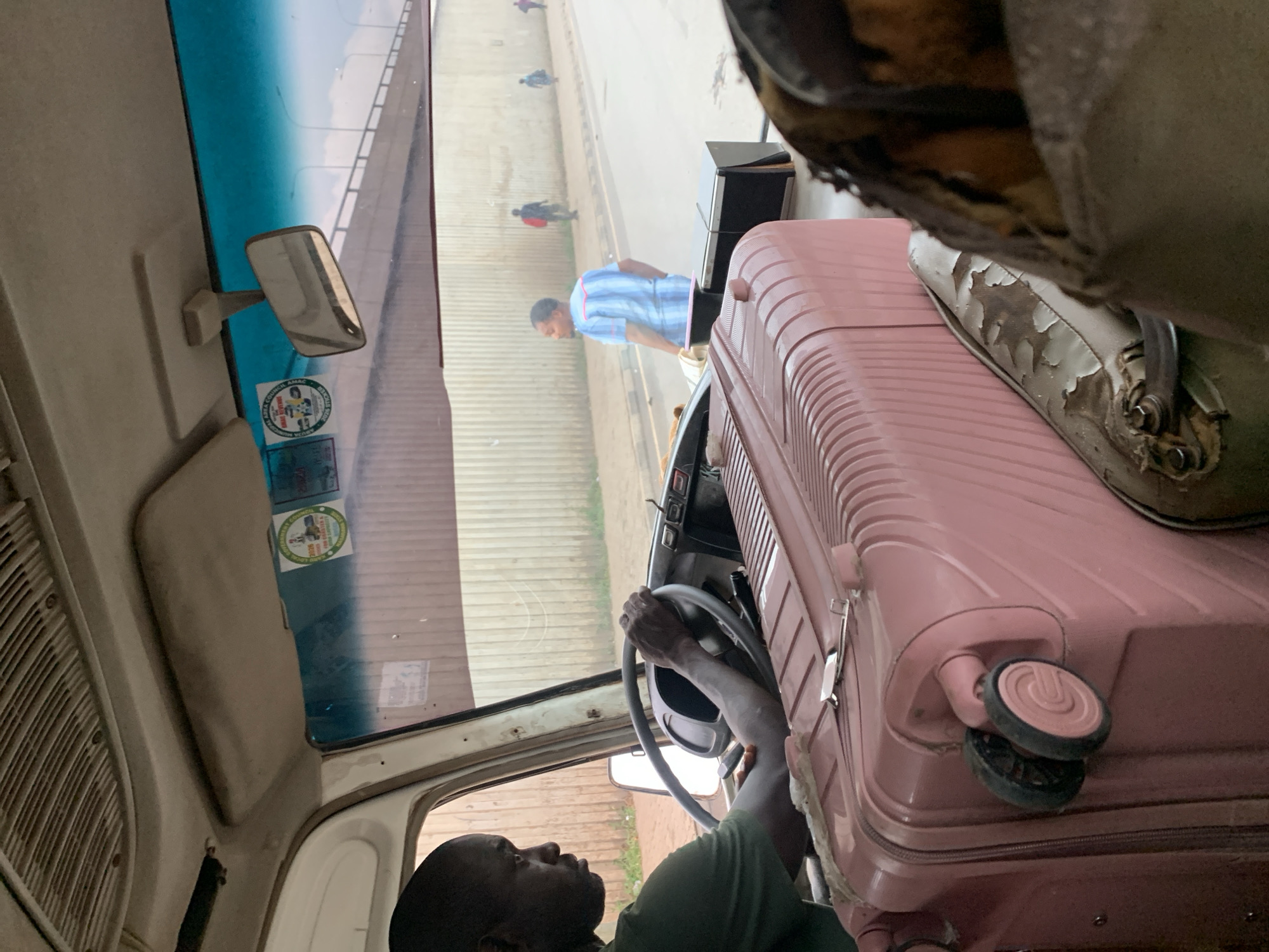 A man in a green t-shirt is driving a bus in the city of Aso, Nigeria. He is holding the steering wheel with both hands and looking forward. A large pink suitcase is positioned in front of him, partially obscuring the dashboard. In the background, a pedestrian wearing a blue and white striped shirt is walking on the sidewalk. The bus is on a road with a concrete wall on the right and a bridge visible in the distance. The sky is overcast.