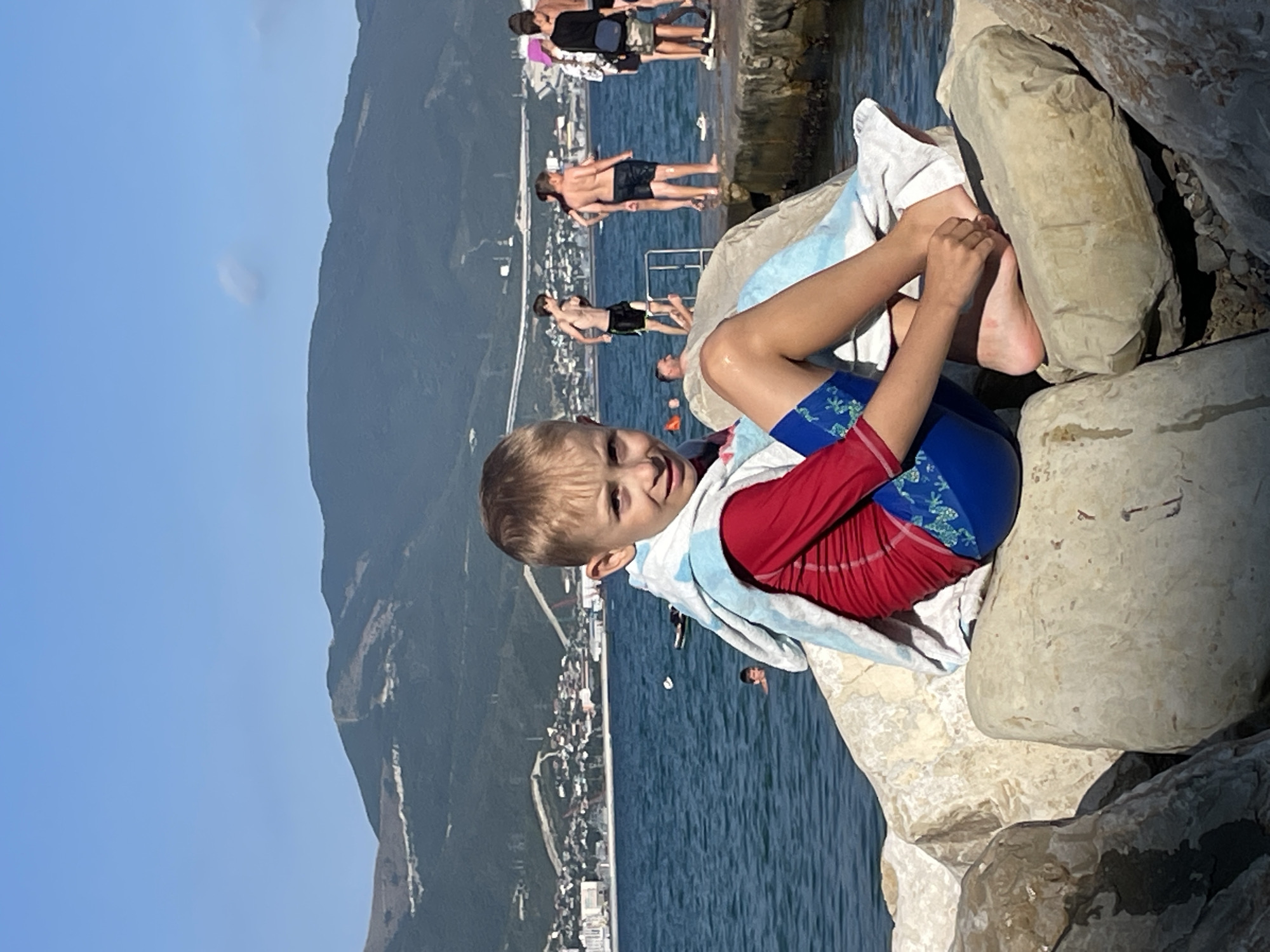 A young boy is resting on rocks near the sea. He is wearing red swimwear and is wrapped in a light blue and white towel. His legs and feet are visible, with one foot tucked under him. In the background, other people are in the water and on a concrete pier. The sea is a deep blue, and a town with buildings is nestled against a green, mountainous landscape. The sky is clear and blue, suggesting it is a sunny day, likely in the afternoon. The overall scene conveys a relaxed atmosphere of a day at the beach or seaside resort in Ol'ginskii, Russia.
