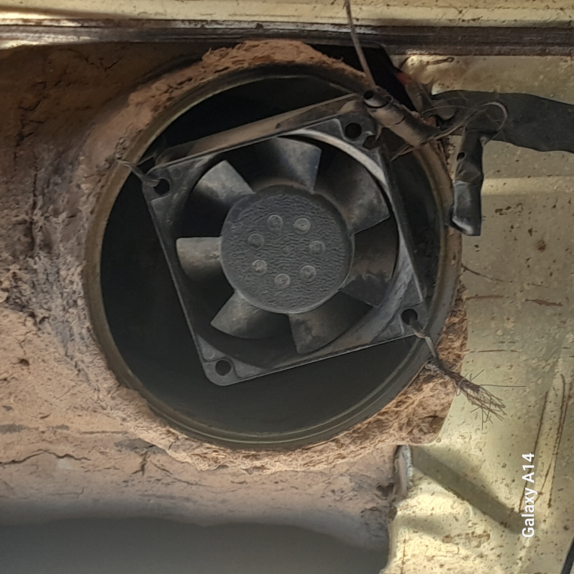The image shows a close-up view of a dusty, black square computer fan installed within a rough, clay-like textured opening. The fan has four blades and a circular center with several small raised bumps. Thin wires, possibly for power, are attached to the fan's mounting bracket on the right side. The surrounding material appears to be a primitive insulation or sealing compound, showing cracks and an uneven, natural texture. Below the fan and opening, a rough, beige-colored surface with some speckling is visible, along with a portion of what appears to be metallic, possibly rusted, structure on the far right. The "Galaxy A14" watermark is present in the lower left corner. The scene is indoors, suggesting an enclosure or housing for the fan, which is likely part of a ventilation or cooling system. The condition of the fan and surrounding materials suggests a utilitarian and possibly aged installation.
