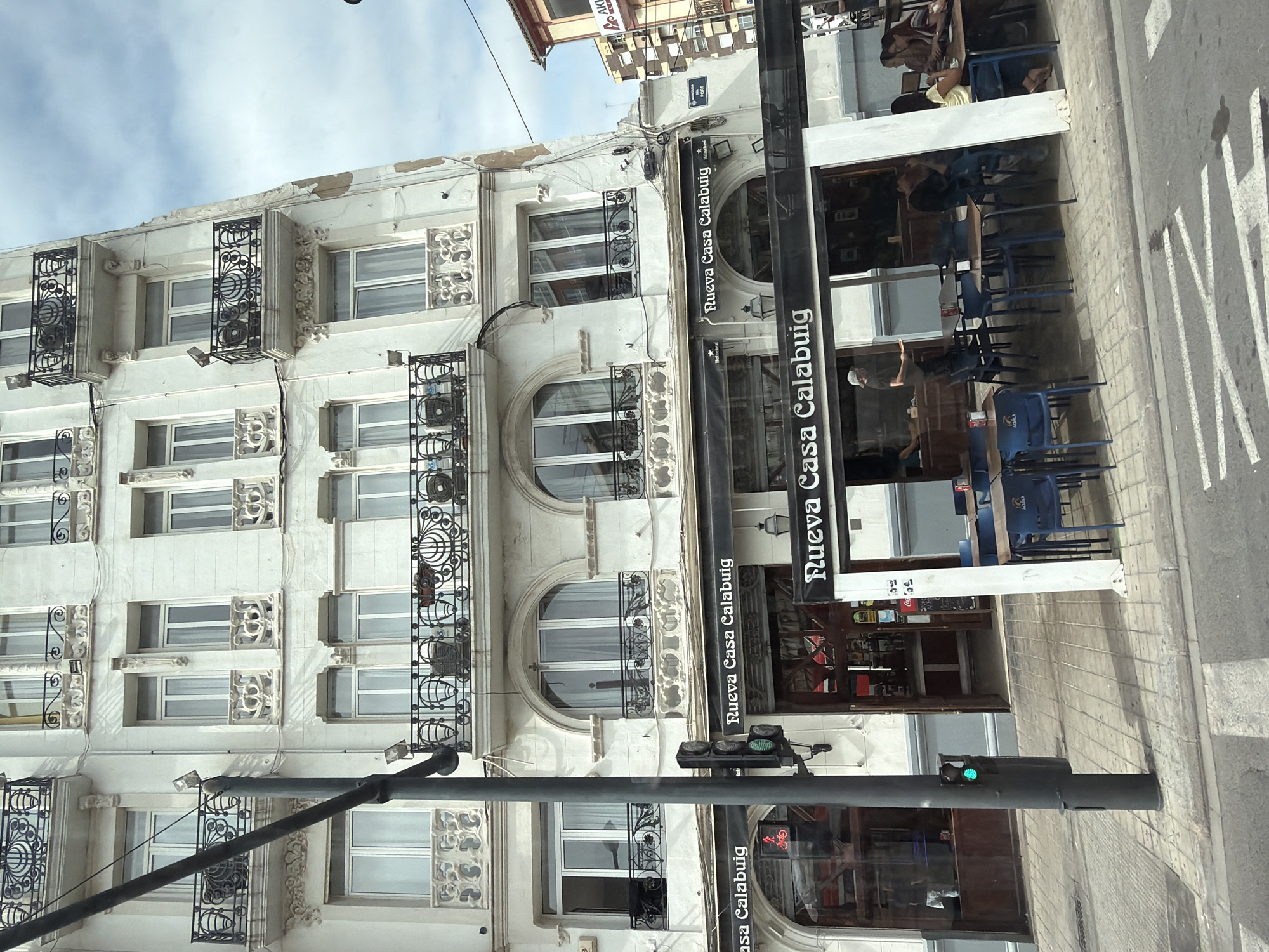 A tall, pale building with ornate balconies and arched windows stands on a city street. A lamppost with a traffic signal is in the foreground. The ground floor of the building features an establishment with awnings that read "Nueva Casa Calabuig." Outside, tables and chairs are set up, with several people seated at them. A pedestrian crossing is visible on the street. The sky is partly cloudy.