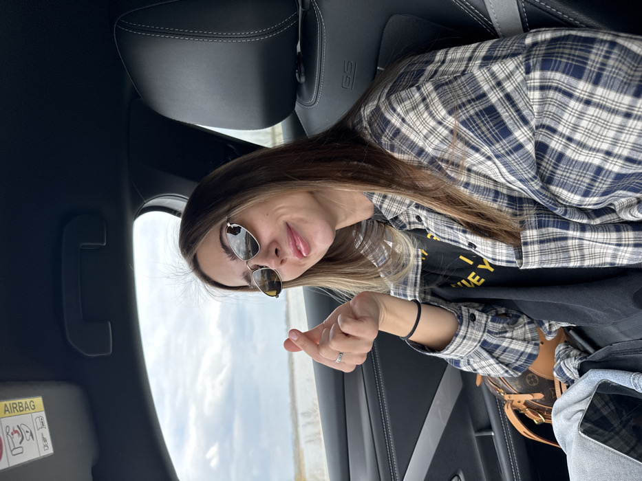 An image displays a young adult female seated in the front passenger seat of a vehicle, looking towards the viewer. She possesses long, light brown hair, wears small, oval-shaped sunglasses with reflective lenses, and her lips are parted slightly in a smile. Her attire consists of a dark t-shirt layered under an unbuttoned blue and white plaid flannel shirt. Her right hand is raised, performing a "finger heart" gesture, with a ring visible on her right ring finger. A black elastic hair tie is on her left wrist.

The vehicle's interior is dark-toned, featuring a black seat with gray stitching and a "GS" logo on the passenger headrest. An "AIRBAG" warning label is present on the sun visor above the subject. Adjacent to her left leg, a small, brown Louis Vuitton handbag with a monogram pattern is visible, alongside a partially obscured dark-colored smartphone and light-colored denim fabric.

Through the vehicle's window on the left, an expansive, flat, light-colored landscape extends to the horizon beneath an overcast sky. This environmental detail is consistent with the geography of Pasohlávky, Czechia, likely depicting one of the Nové Mlýny reservoirs or a wide, open plain, possibly covered in snow or ice, with a faint line of bare vegetation visible in the distance.