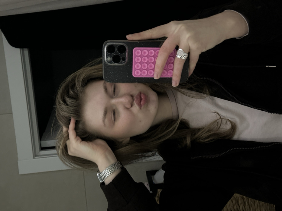 An indoor mirror selfie depicts a single female subject in Kazan, Russia. The subject, with light brown hair, is positioned in the foreground, facing the camera's reflection. She is wearing a black zippered hoodie over a light-colored crew-neck shirt. Her left hand is raised, fingers interacting with the hair near her scalp. A silver-toned metal watch with a link bracelet is on her left wrist. Her right hand holds a dark-colored smartphone, equipped with a speckled case and a rectangular pink accessory featuring multiple circular tactile buttons. A large, ornate silver-toned ring is present on her right ring finger. The subject's facial expression involves puckered lips and partially closed eyes. The background, reflected in the mirror, consists of a light-colored wall section and a white doorframe on the left, with a predominantly dark, featureless area on the right. A woven texture and a blurry white object are partially visible in the lower-left background.