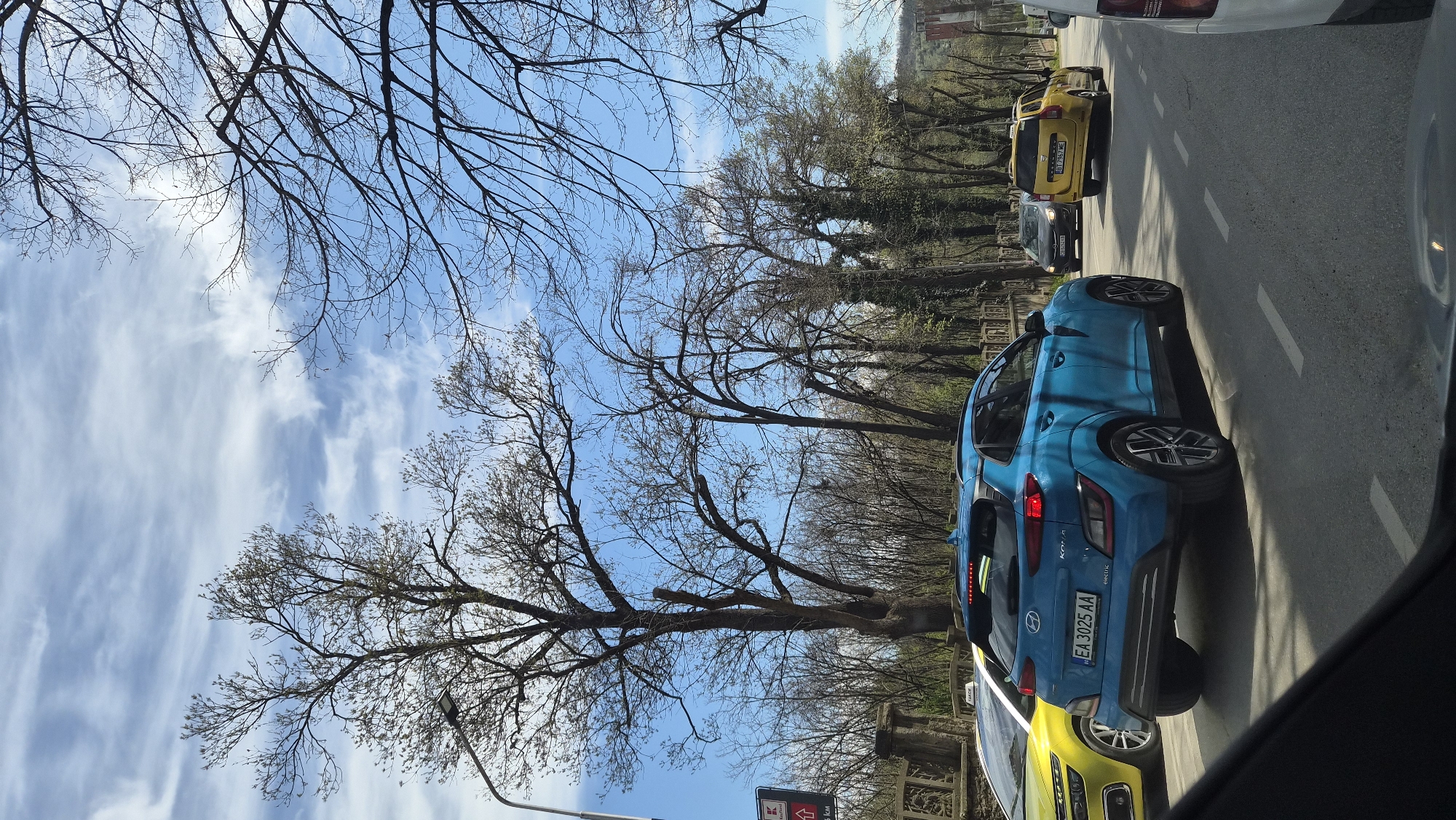 The image captures a street scene in an urban environment. A blue car, a Hyundai Kona Electric, is prominently featured, partially obscured by a yellow car. The blue car's license plate reads "EA 3025 AA." Behind the blue and yellow cars, other vehicles are visible, including a yellow Dacia. A row of trees, largely bare of leaves, lines the side of the road. The sky is visible through the branches, featuring scattered clouds and a blue hue. A sign is visible in the lower left corner, near a brick structure and a street light. The image is taken from inside a car, with the edge of the car door visible in the foreground.