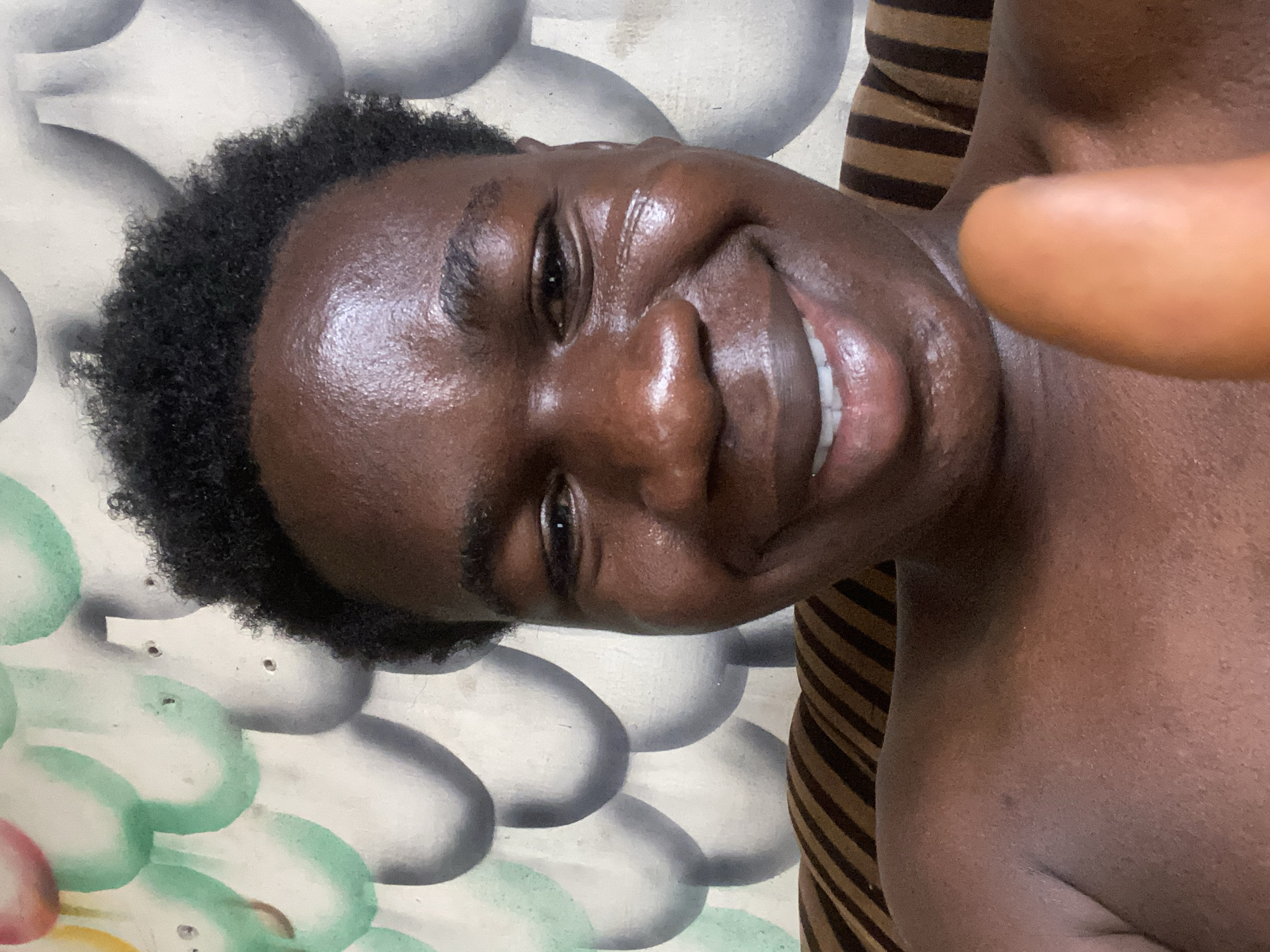 A close-up selfie of a dark-skinned person smiling broadly. The person has short, curly black hair and is shirtless. The background consists of a patterned wall and a striped fabric. The location in Mararaba, Nigeria, is not discernible from the image.