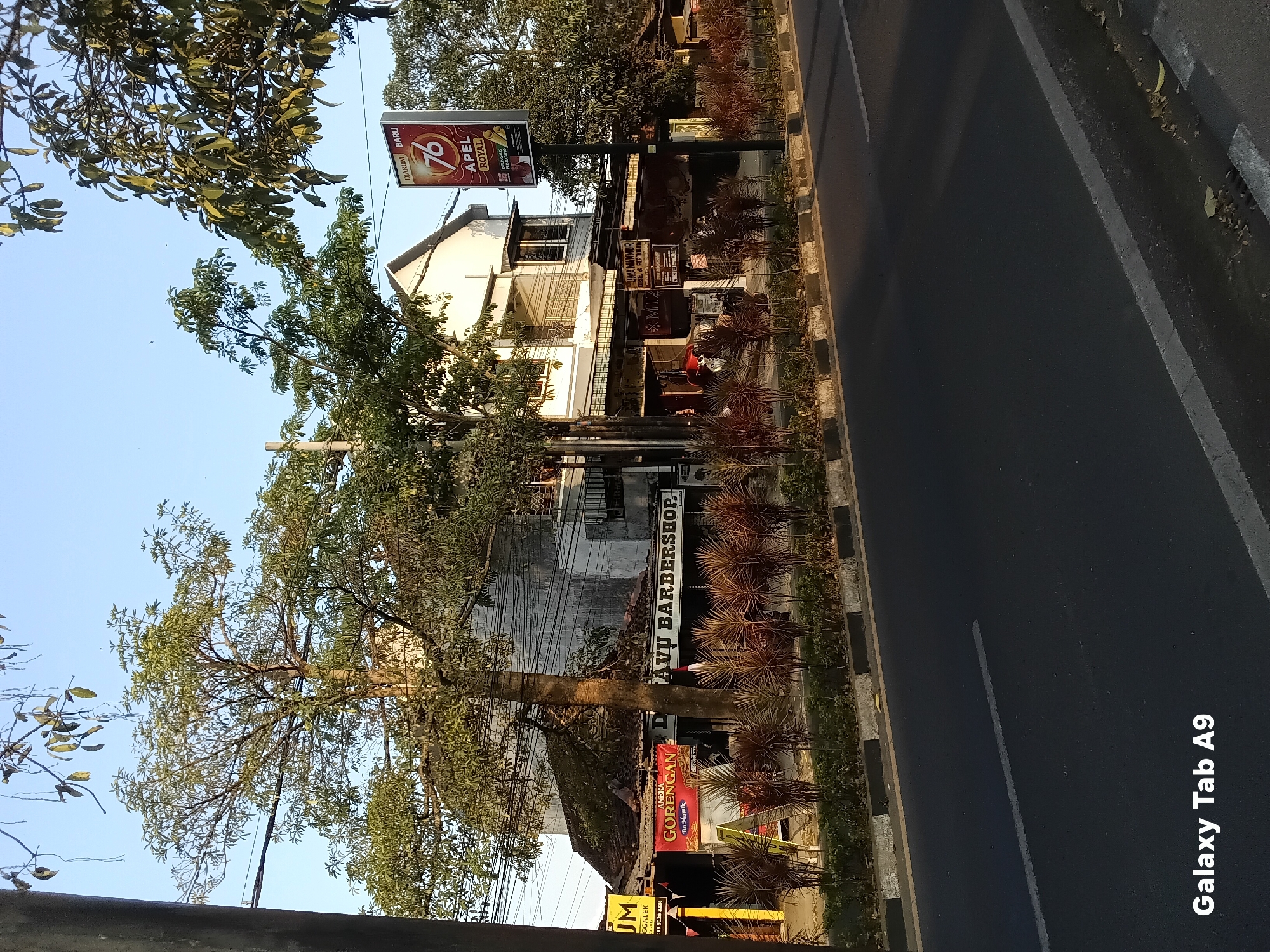 The image shows a street scene in Kelutan, Indonesia, with a focus on a row of shops and buildings along the road. The buildings appear to be a mix of older structures with tiled roofs and a more modern, multi-story building. In front of the shops, there is a sidewalk area with decorative plants and a road in the foreground.
On the left side of the image, a sign with the text "UM" and "GGALEK" is visible, suggesting a business name and location. Next to it, a red banner with the text "ANEKA GORENGAN" and "Bu Manik" indicates a food vendor selling fried snacks. Further down the row of shops, a sign reads "DAVU BARBERSHOP," identifying a barbershop.
A tall, vertical billboard stands on the right side of the road, featuring an advertisement for "76 APEL ROYAL," with images of apples and a person. Trees are present throughout the scene, with one large tree prominently positioned in front of the buildings. Overhead, electrical wires are strung across the scene. The sky is clear and blue, indicating fair weather. The overall atmosphere suggests a typical urban street in Indonesia during daylight hours. A watermark in the lower left corner of the image reads "Galaxy Tab A9."