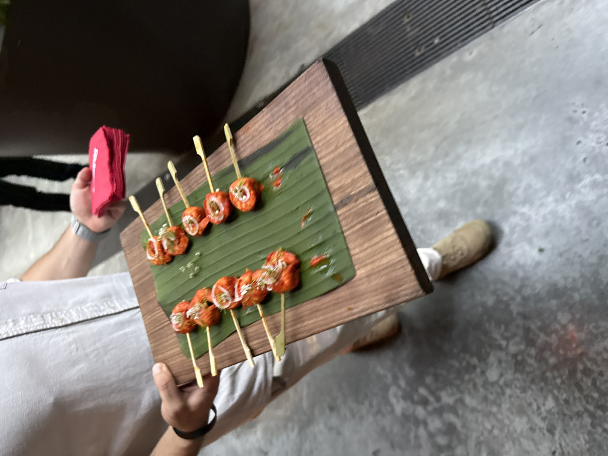 An unseen person holds a wooden board with skewers of food. The food appears to be shrimp or some other seafood, coated in a red sauce and topped with white and green garnishes. The skewers are placed on a banana leaf atop the wooden board. The person is wearing a light-colored shirt and tan pants, and they are holding a stack of red napkins in their other hand. The background is blurred but suggests an outdoor or semi-outdoor setting with a concrete floor and a dark, textured surface. The image is taken from a slightly high angle, looking down at the board and food.