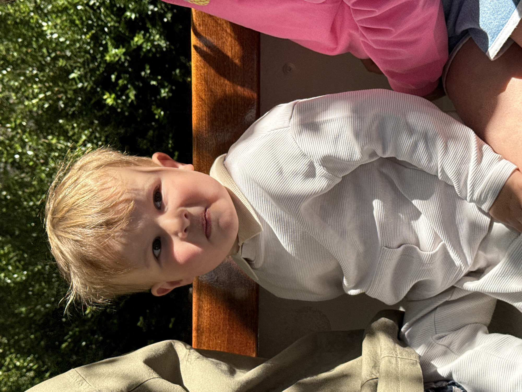 A close-up, medium-shot photograph shows a young child with blond hair and light skin wearing a striped white shirt. The child is seated at an outdoor location. Behind the child is a dark-colored wooden railing. To the left is lush green foliage that appears to be from trees. In the upper portion of the image, parts of clothing and limbs from other individuals are visible. There is a pink sweatshirt and blue denim. The surroundings suggest an outdoor, possibly garden-adjacent setting. There is no information from which to specifically determine that the image was taken in the city of Bruges, Belgium.