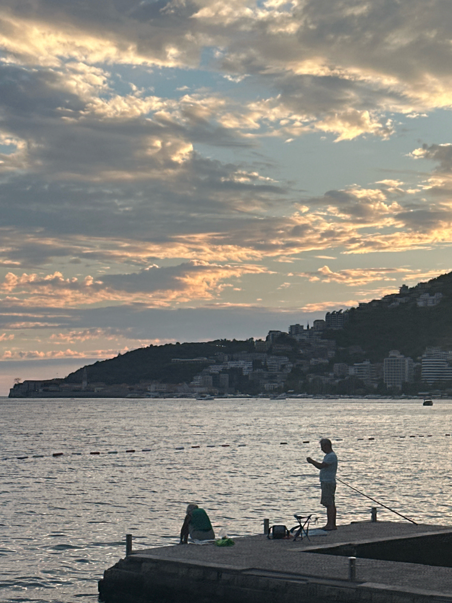 This is a photograph taken from a pier looking towards the coast of Budva, Montenegro at sunset. Two individuals are visible on the pier. In the foreground to the left, a person is hunched over, seemingly engaged in an activity on the pier. To the right, a standing individual is holding a small object, possibly a phone, and appears to be looking at it. A fishing rod extends from the pier into the water near the standing person. A foldable chair and a bag are also on the pier. The sea is calm with a few buoys marking a boundary in the water. In the background, a densely populated hillside with buildings and structures overlooks the coastline. The sky is filled with dramatic clouds illuminated by the warm, soft light of the setting sun, casting a pastel glow across the scene.