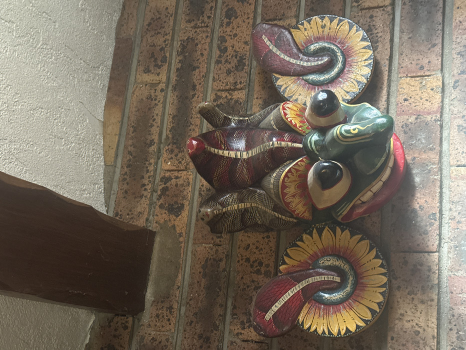 The image displays a multi-colored carved wooden mask mounted on a reddish-brown brick wall. The mask features a central green, stylized reptilian or amphibian face with prominent black-eyed protrusions, a wide red-lined mouth, and white teeth details. Above the face, a headdress features five visible stylized cobra heads with reddish-brown and grey striped patterns, the central one tipped in red. Two large, ornate circular elements, resembling stylized sunflowers or coiled shells in yellow, red, and green, extend from either side of the central face. The brick wall features dark mortar lines. In the upper left quadrant, a dark brown, curved wooden beam extends from an off-white textured ceiling surface, partially obscuring the top of the brickwork. No individuals or active interactions are depicted. The scene is located in Torcy, France.
