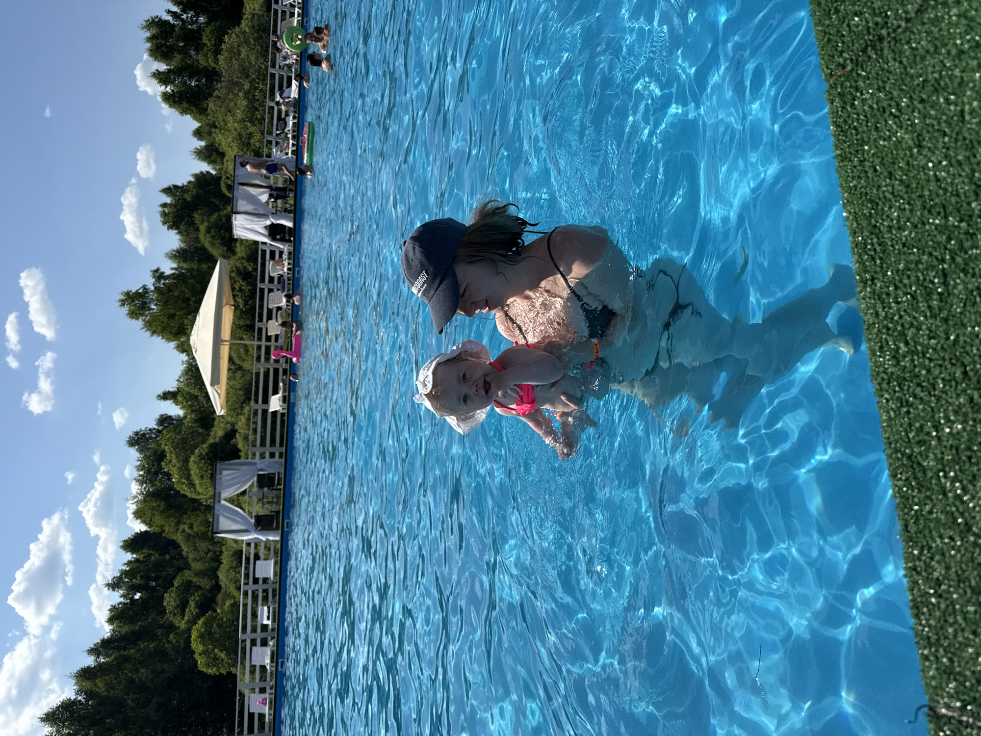 In the city of Koloksha, Russia, a woman wearing a black bikini top and a dark baseball cap holds a baby in a swimming pool. The baby is wearing a pink swimsuit and a white sun hat. They are both in the bright blue water, which reflects the sunlight. In the background, other people are swimming and relaxing by the pool, with trees and a cloudy blue sky visible. Some lounge chairs and cabanas are set up around the pool.