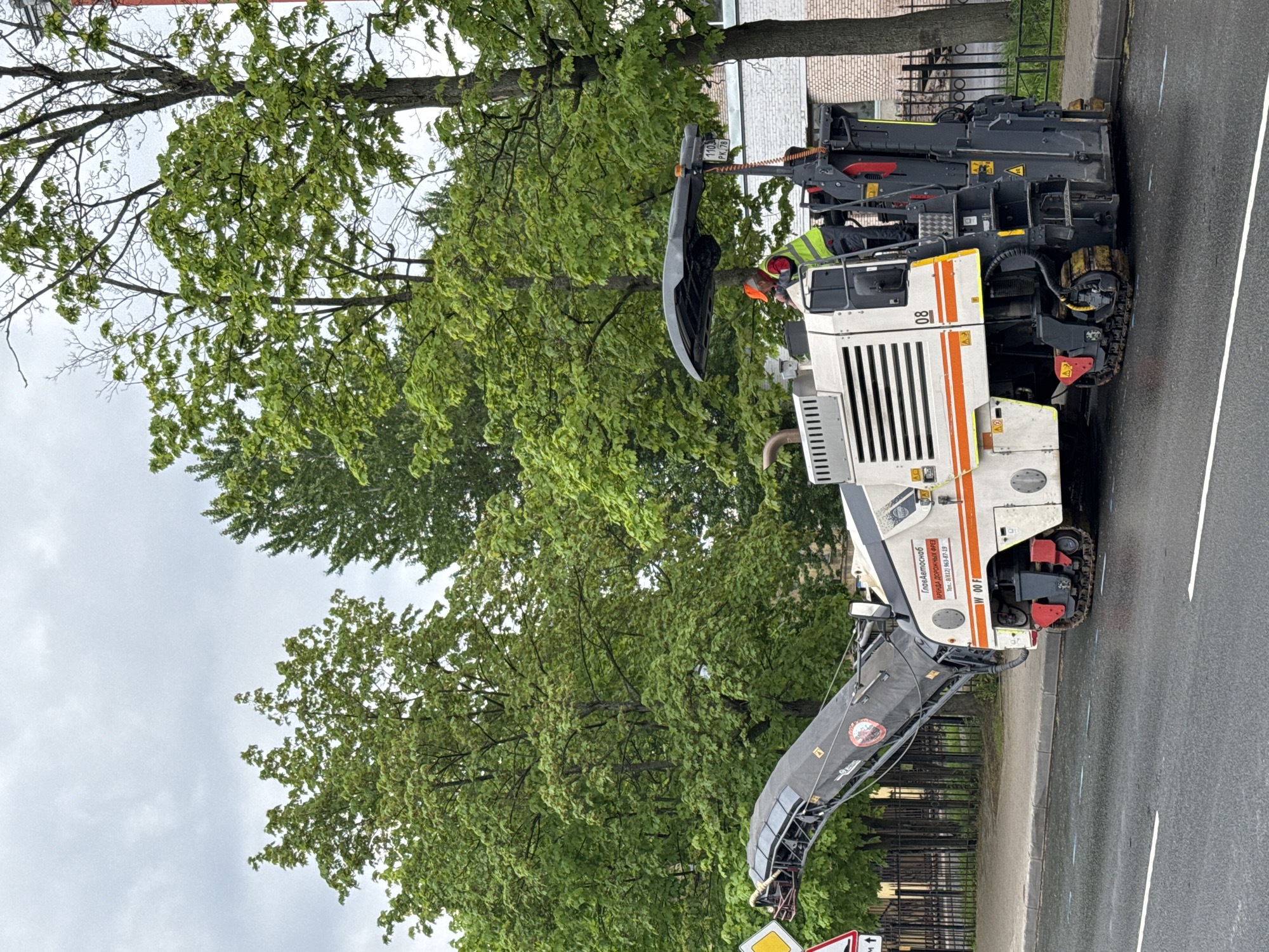 Here is a detailed description of the image:
**Overall Scene:**
The image shows a street scene in what appears to be Saint Petersburg, Russia, indicated by the Cyrillic writing on the machinery. It's an urban environment with mature trees and a mix of older and newer buildings in the background. The focus is on road maintenance equipment.
**Objects:**
1. **Road Milling Machine**: A large, heavy-duty road milling machine is the central object. It is white with orange stripes and appears to be in operation, likely removing the top layer of asphalt.
2. **Trees**: Lush, green trees line the street, giving a sense of an established urban area.
3. **Road**: A paved road, likely asphalt, is visible with some markings and possible evidence of recent rain or dampness.
4. **Building**: Brick building is in the background.
5. **Fence**: Wrought iron fences can be seen in sections.
**People:**
1. **Worker**: A worker is present on the milling machine, wearing a safety vest and a hat.
**Actions:**
The road milling machine appears to be in the process of milling the road surface. The worker seems to be operating or overseeing the machinery.
**Environment:**
The weather appears overcast. The presence of trees suggests it is either spring or summer. The setting is urban with both greenery and infrastructure.