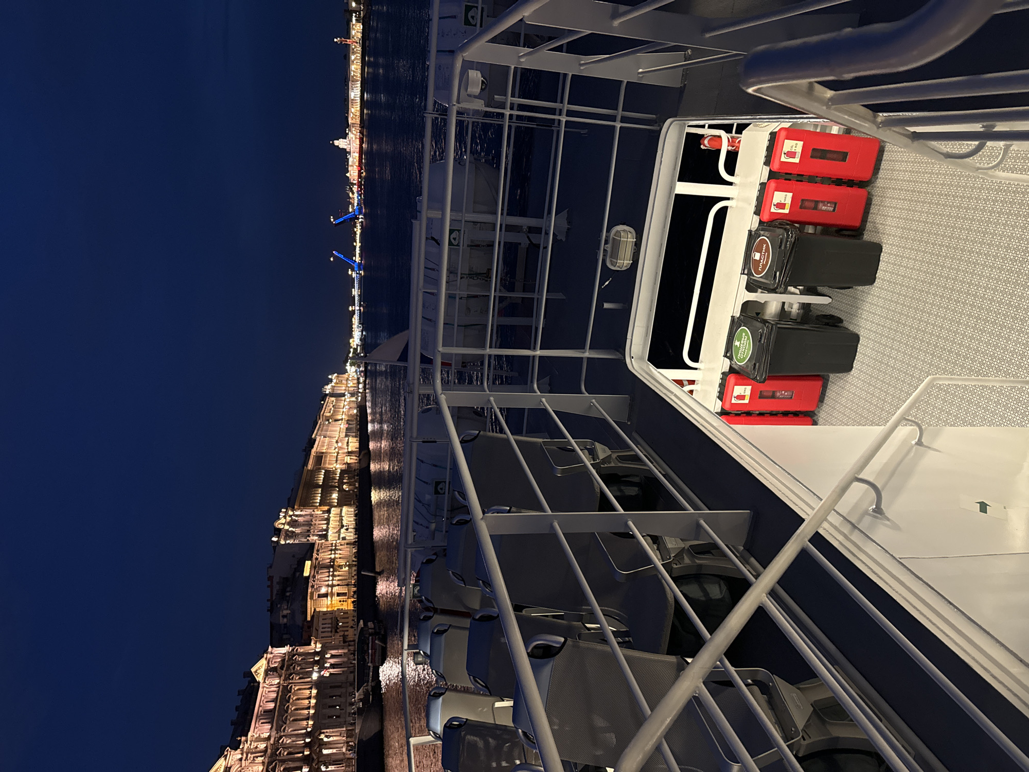 A night view from a boat on the Neva River in Saint Petersburg, Russia, looking towards the Palace Embankment. The waterfront is illuminated with the warm lights of historic buildings, including what appears to be the General Staff Building and the Winter Palace. In the distance, a drawbridge is raised and lit with blue lights. The foreground of the image shows the deck of the boat, with rows of empty, dark-colored seats facing the river. White railings and metal structures are visible throughout the deck. A set of stairs leads down from the main deck. On the lower deck, there are two black trash receptacles labeled "ПРИИМИТЕ ОТХОДЫ" (Please take waste) and "ПЛАСТИК" (Plastic). Next to them are two red fire extinguishers. The sky is a deep, clear blue, indicating nighttime.