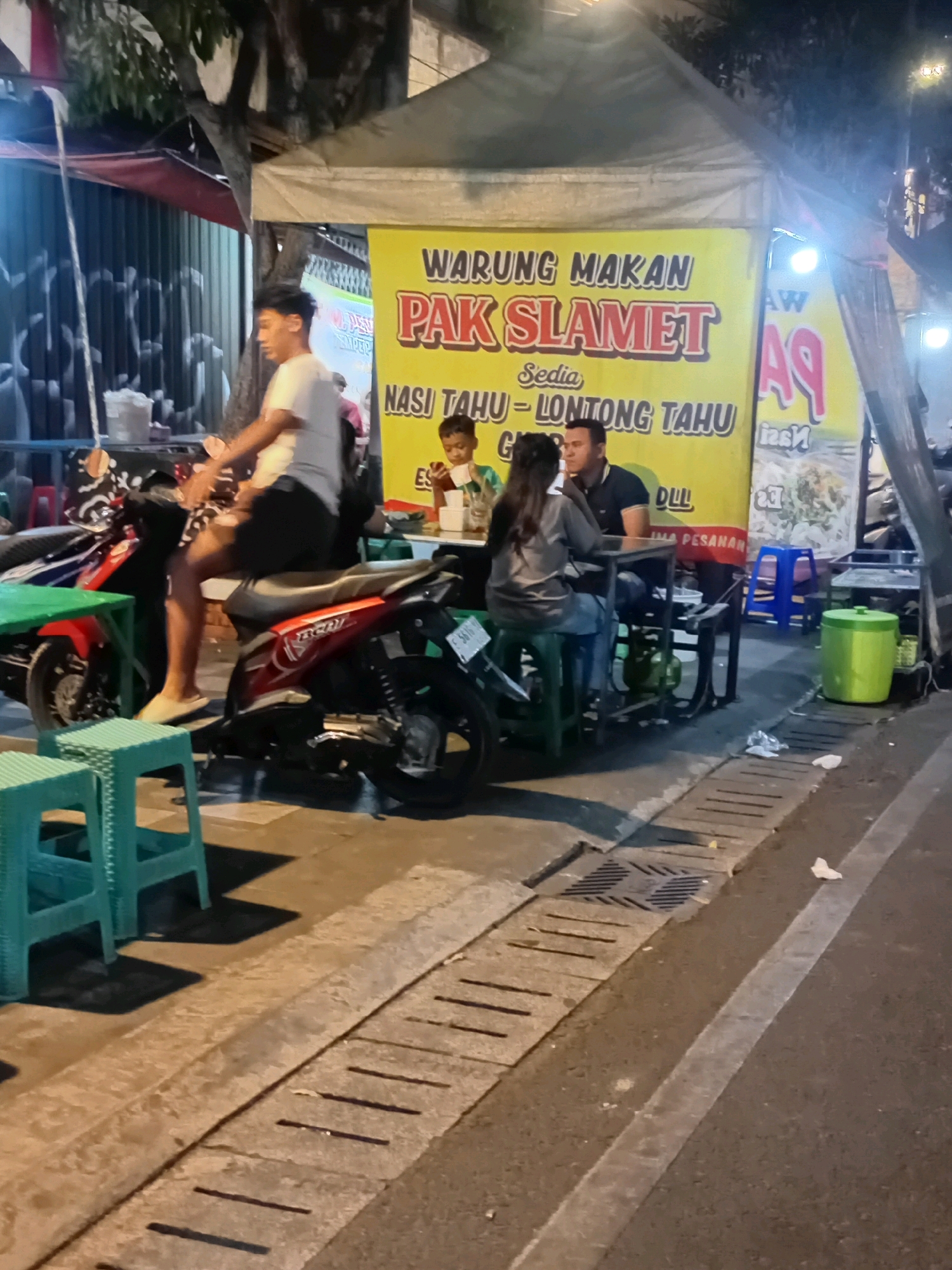 The image depicts a street scene at night, likely in Kudus, Indonesia, featuring a food stall with a yellow banner advertising "WARUNG MAKAN PAK SLAMET". The banner also lists menu items such as "NASI TAHU - LONTONG TAHU". Several individuals are present. A young man in a white t-shirt and black shorts is seated on a red motorcycle in the foreground. Behind him, a boy is seated at a small table. To the right of the boy, a man and a woman are seated at another table, seemingly interacting. The stall is set up under a tent, with various items visible, including tables, chairs, and a green trash bin. The surrounding environment appears to be a sidewalk and a street, with drainage grates present. The lighting suggests it is nighttime, with artificial lights illuminating the area.