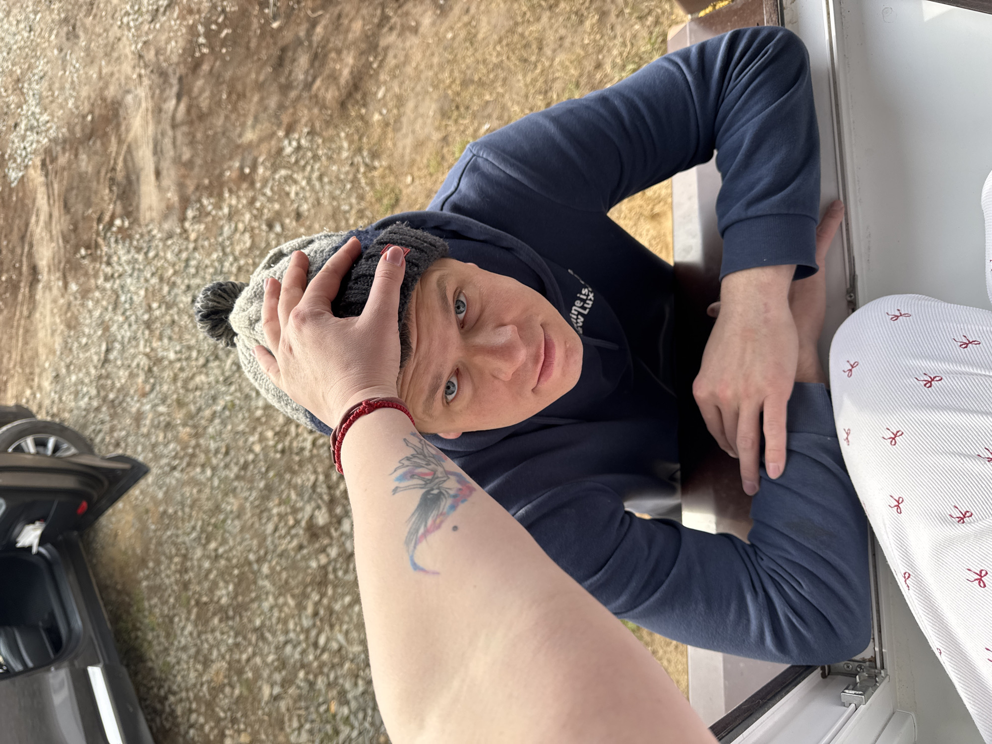 A high-angle view photograph depicts a man wearing a dark blue hooded sweatshirt and a gray knit hat with a pompom. A person's arm with a visible tattoo and red string bracelet is partially visible, with the hand touching the man's hat. The man is looking up at the camera. He is positioned in an area that appears to be the edge of a window frame. Outside, there is a ground surface covered in gravel, with a dark-colored car visible in the lower left corner. A white fabric with red bow patterns is visible on the right side of the frame. The photo was taken outdoors in Gornyi (Dvurechenskoe S/P), Russia.