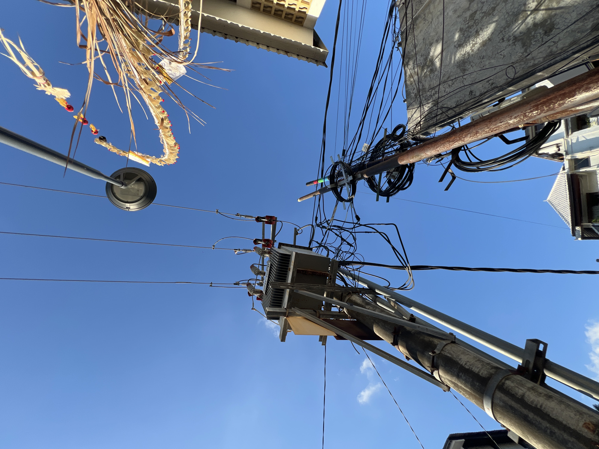 The image, taken from a low angle, shows a dense network of utility poles and wires against a clear blue sky in North Kuta, Indonesia. The poles are primarily concrete and metal, with one appearing older and more weathered. The poles support an electrical transformer and a complex web of power and communication cables. A lamp post is visible in the left foreground, adorned with a traditional Balinese offering (canang sari). Buildings are partially visible at the top of the frame, indicating an urban or suburban setting.