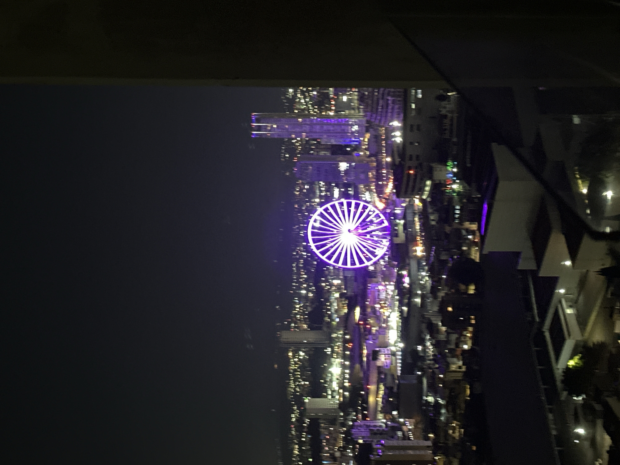 The image displays a high-angle, panoramic nighttime view of the city of Puebla, Mexico. Centrally located in the midground is the Estrella de Puebla, a large Ferris wheel brightly illuminated with purple and white lights, forming a radial spoke pattern. To the right of the Ferris wheel, a prominent, tall skyscraper is vertically illuminated with purple and white lights. The surrounding urban landscape is densely populated with numerous buildings of various heights, many displaying illuminated windows and outlines in white, yellow, and purple hues. Thousands of smaller, blurred lights are distributed throughout the cityscape, consistent with streetlights and vehicle traffic. The foreground on the right side of the frame is occupied by a dark, vertical structural element, likely part of a building or balcony, partially obscuring the lower-right portion of the scene. Below this structure, dark building rooftops and a few ground-level lights are visible. The sky above is dark and clear, characteristic of an urban night sky. No individuals or discernible actions are present.