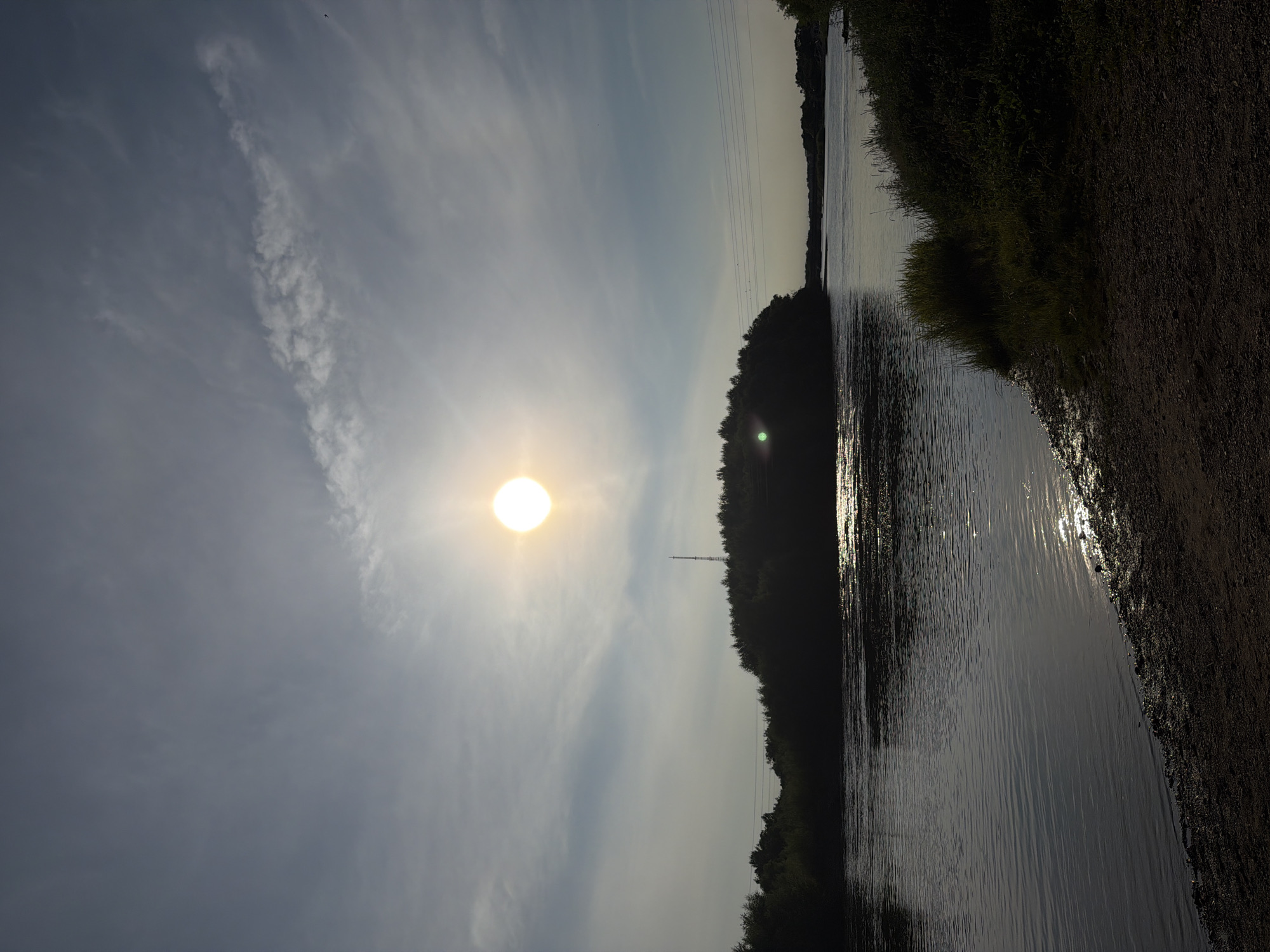 The image depicts a serene natural landscape, likely a riverbank, under a bright, partially obscured sun. The sun, a glowing orb, is positioned in the upper portion of the frame, casting a brilliant light that creates a luminous haze in the sky. Wispy, light-colored clouds are scattered across the expanse of the sky.
Dominating the mid-ground is a dense, dark silhouette of trees forming an island or peninsula in the water. A tall, slender tower, possibly a radio or transmission tower, is visible rising from the treeline on this landmass. Several power lines, suspended between unseen poles, stretch horizontally across the upper mid-ground.
The foreground is composed of a body of water, appearing calm with gentle ripples. The sunlight reflects off the water's surface, creating shimmering patterns. The bank of the river is visible in the lower portion of the image, covered with gravel and sparse vegetation, with more dense foliage visible on the right side of the frame. The overall impression is one of tranquility and natural beauty at dusk or dawn. There are no people visible in the image.