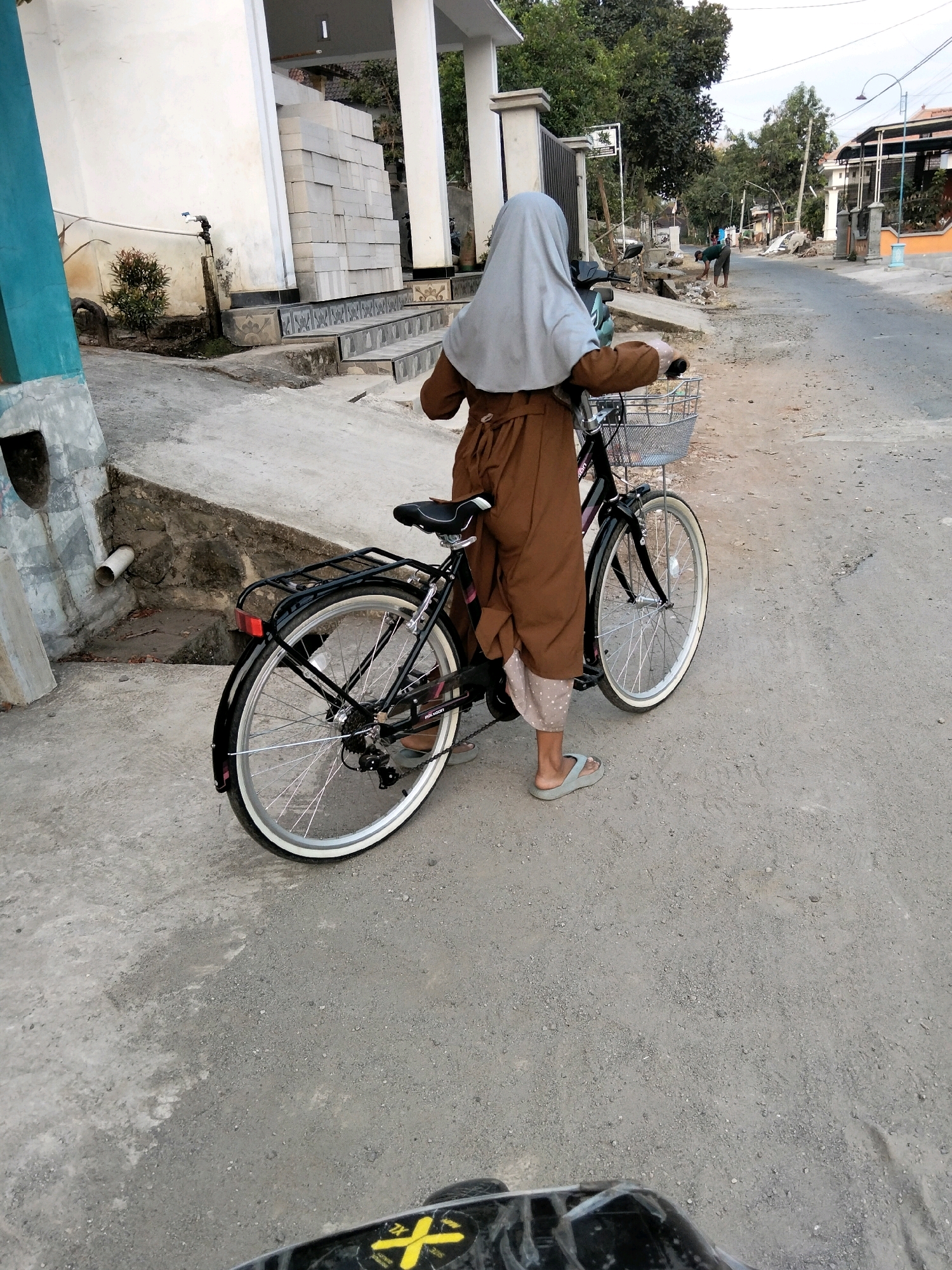 In Jenangan, Indonesia, a person wearing a brown robe and a light grey hijab stands next to a black bicycle with a wire basket on the handlebars and a rear rack. The person is facing away from the camera, with their right hand on the bicycle's handlebar and their left hand on the seat. They are wearing light blue flip-flops. The bicycle has a black frame, a black seat, and silver spokes with white-rimmed tires. The scene takes place on a paved street with some cracks and gravel. To the left of the bicycle, there is a concrete structure with a drainpipe. In the background, there are buildings, trees, and other people in the distance. One person is working on the side of the road. The sky is overcast.