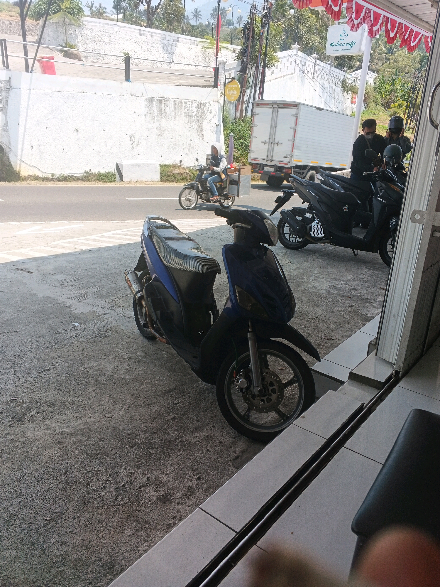 In Pakopen, Indonesia, this image captures a roadside scene with multiple motorcycles and people. In the foreground, a blue scooter is parked on a concrete surface, its front wheel and handlebars visible. Behind it, two black motorcycles are also parked, one with a white honeycomb pattern on its side. Three individuals are present: two men wearing black shirts and dark headwear, one of whom appears to be talking on a phone, and a woman wearing a light-colored hooded jacket and jeans, riding a motorcycle in the background. A white truck is visible on the road, which is paved and marked with white lines. The background features a white wall, some trees, and a white sign with red and white striped banners overhead. The overall setting appears to be an outdoor establishment, possibly a cafe, with a tiled floor and metal railings.