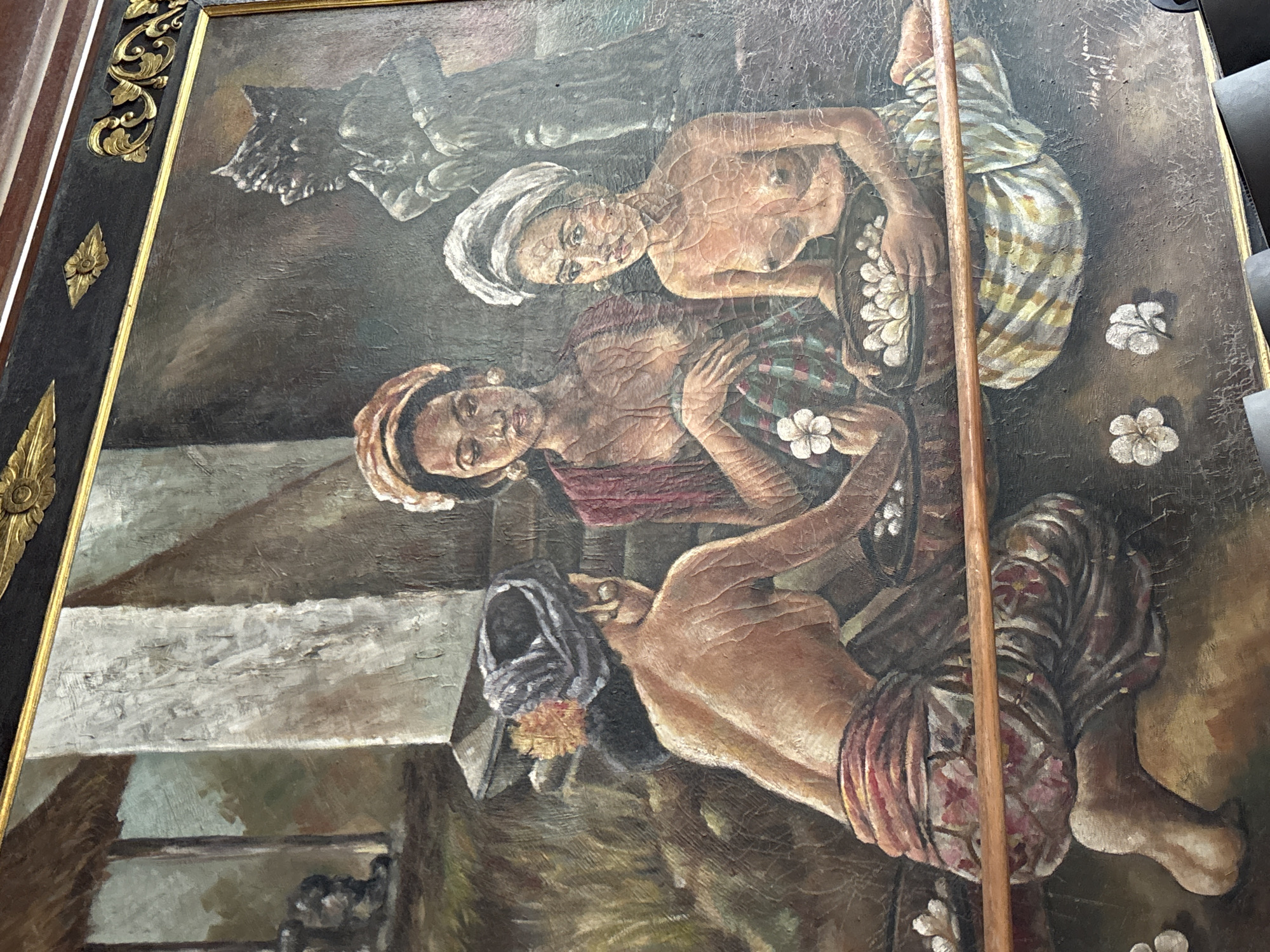 The image shows an oil painting with a thick, ornate dark wood and gold frame. The painting depicts three semi-nude Indonesian women, rendered in a realistic, somewhat aged style. One woman is lying on her back, facing the viewer, with a white cloth wrapped around her head and a striped garment around her lower body. The second woman is seated, also facing the viewer, wearing a headdress and a maroon upper garment. The third woman is reclining, with a dark head covering. All three women are positioned around a basket of white flowers. The environment includes a dark, indistinct background with what appears to be steps or levels in the lower left corner, and a grey, sculptural element above the women. Loose white flowers are scattered near the women. The scene is likely set in Indonesia, possibly based on the style of the women's garments and features. Pekutatan, Indonesia is mentioned as a location, which provides a cultural context.