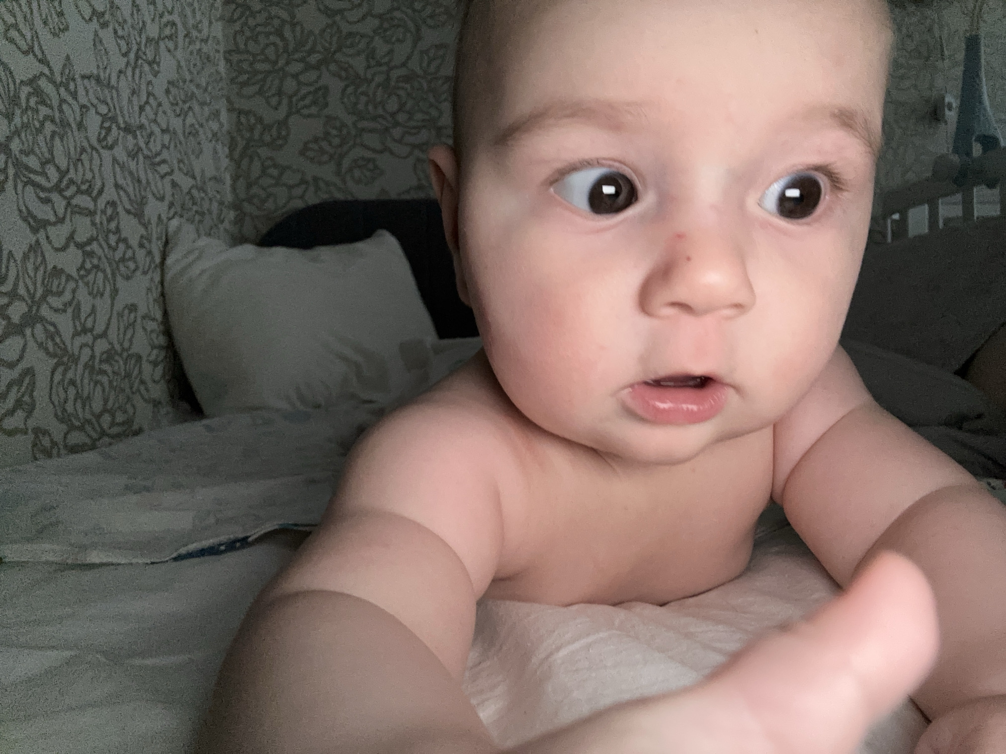 A close-up, slightly angled photo shows a baby lying on a bed. The baby is shirtless, with light skin, brown hair, and brown eyes that appear wide and expressive. The baby is propped up on their forearms, with one hand more prominently in the foreground, partially out of focus. The baby's mouth is slightly open. Behind the baby is a bed with a white pillow and a patterned wallpaper. The photo was taken indoors, likely in a home. While the photo was taken in the city of Chaikovsky, Russia, there is nothing identifiable that would suggest this location in the image.