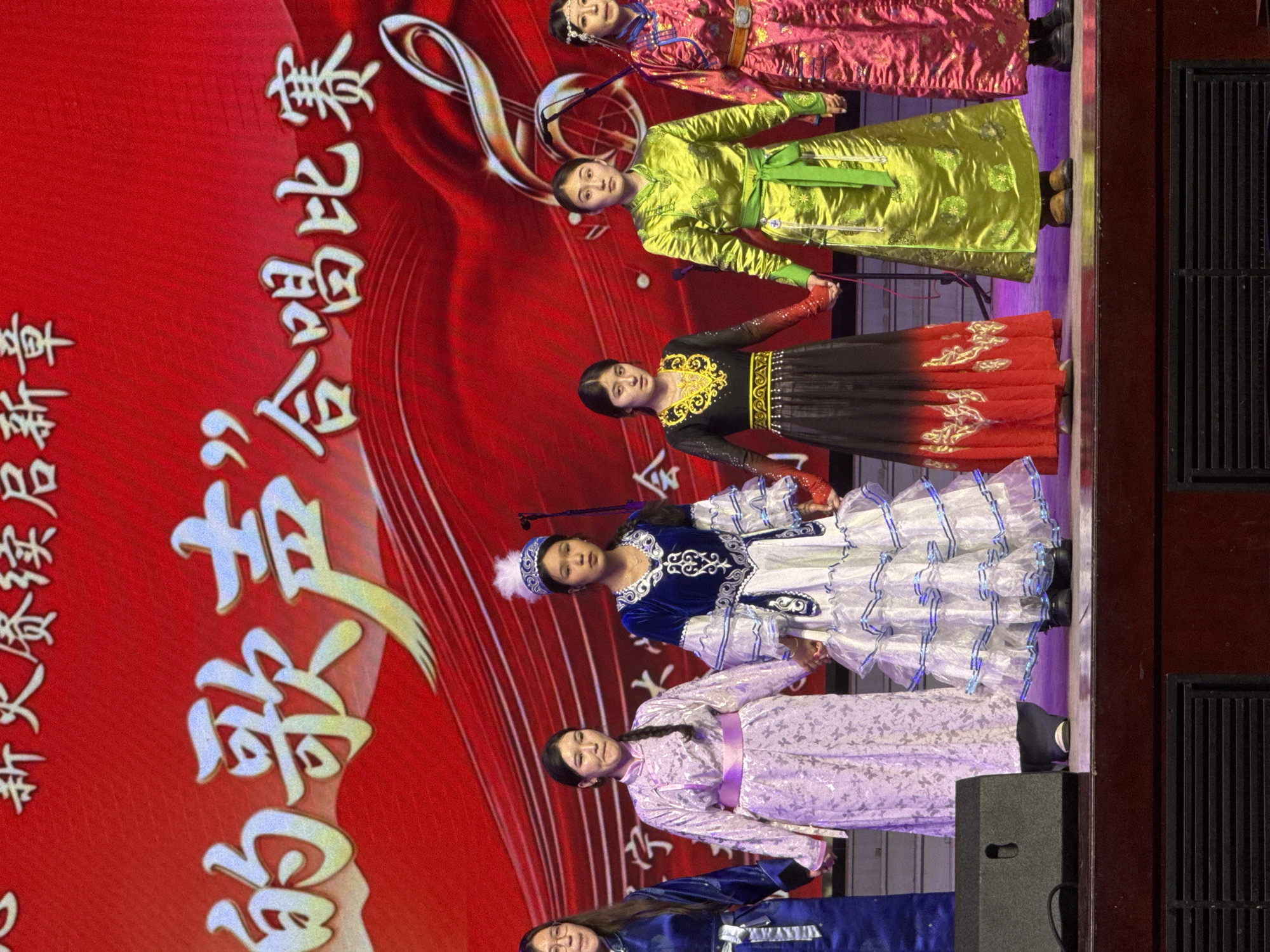 Six women in traditional attire are performing or posing on an indoor stage. The stage features a red backdrop adorned with large Chinese characters and musical notes, including a prominent golden treble clef. The women are arranged in a line, holding hands, facing forward. Each woman wears a distinct, colorful long dress with intricate patterns and details, some with sashes or headpieces, suggesting diverse cultural representation.
From right to left, the costumes include: a pink patterned dress with a dark trim, a bright lime green robe with a lighter green sash and gold embroidery, a black and red ombré dress with gold embroidery on the bodice and skirt, a blue top with white embroidery paired with a voluminous white and blue patterned skirt and a white feathered hat, a light purple patterned dress with a pink sash, and a partially visible dark blue dress with white patterns. Some women are holding microphones on stands, indicating a singing performance. The stage floor is dark, with a purple glow under the performers' feet, and a speaker is visible at the front right edge.
Visible Chinese text on the red backdrop, when read vertically from top to bottom, includes "合唱比赛" (Chorus Competition) as the main title, and "新火赞颂启新章" (New Fire Praise Opens a New Chapter) as a smaller thematic subtitle on the left. The women generally have neutral to slightly smiling expressions, looking towards an unseen audience or camera. The setting is a cultural event, likely a competition or performance, in Guta, China.