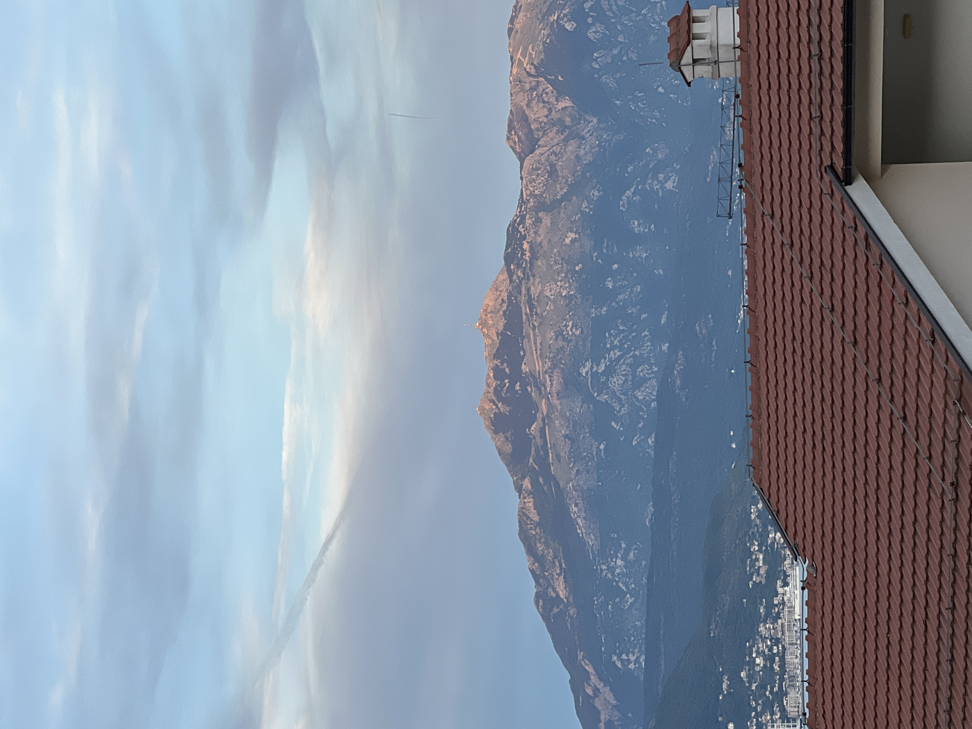 The image captures a mountain range illuminated by the golden light of sunset, viewed from a residential area in Baošići, Montenegro. In the foreground, a red-tiled roof dominates, with a white chimney structure and part of a building visible. Beyond the roof, a coastal town with scattered buildings nestles against a hilly landscape, leading to the expansive, rugged mountains in the distance. The sky above is a soft blend of blue and grey, with wispy clouds and two faint contrails stretching across it. The overall scene evokes a peaceful evening atmosphere overlooking the Adriatic coast.