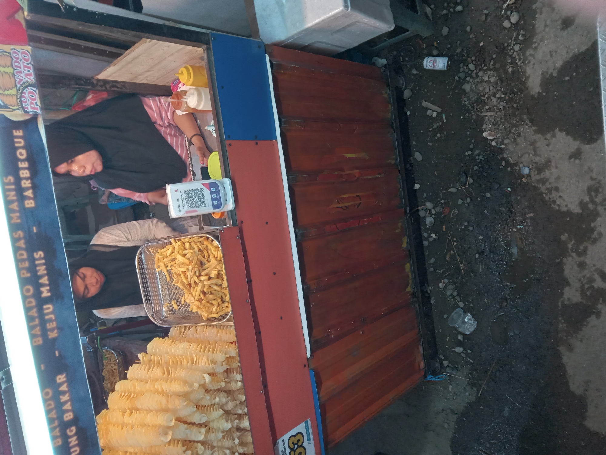 A street vendor's stall displays tornado potatoes and french fries. Two women, wearing headscarves, are working at the stall. The stall is decorated with colorful signage that includes text in Indonesian. The ground around the stall is littered with debris, including small rocks and what appears to be a discarded can and bottle cap. A QR code is visible on the counter, likely for payment. The lighting suggests it is either daytime or under bright artificial light.
