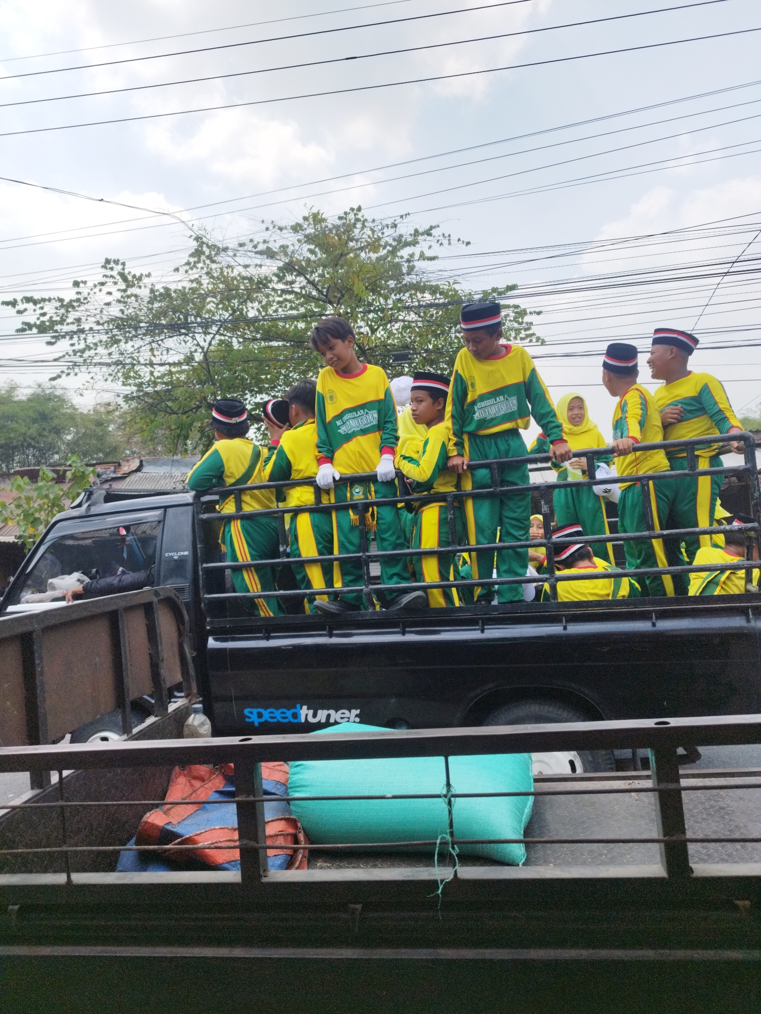 In Bumirejo, Indonesia, a black pickup truck carries a group of young people wearing matching green and yellow uniforms with black hats adorned with red and white stripes. They appear to be participating in a procession or event, standing and interacting with each other on the truck bed. The truck is parked on an unpaved surface, and behind it, a tree with green foliage and power lines are visible against a cloudy sky. In the bed of the truck, there are also some bundled items, including what appear to be large sacks or tarps. The truck has "speedtuner." written on its side.