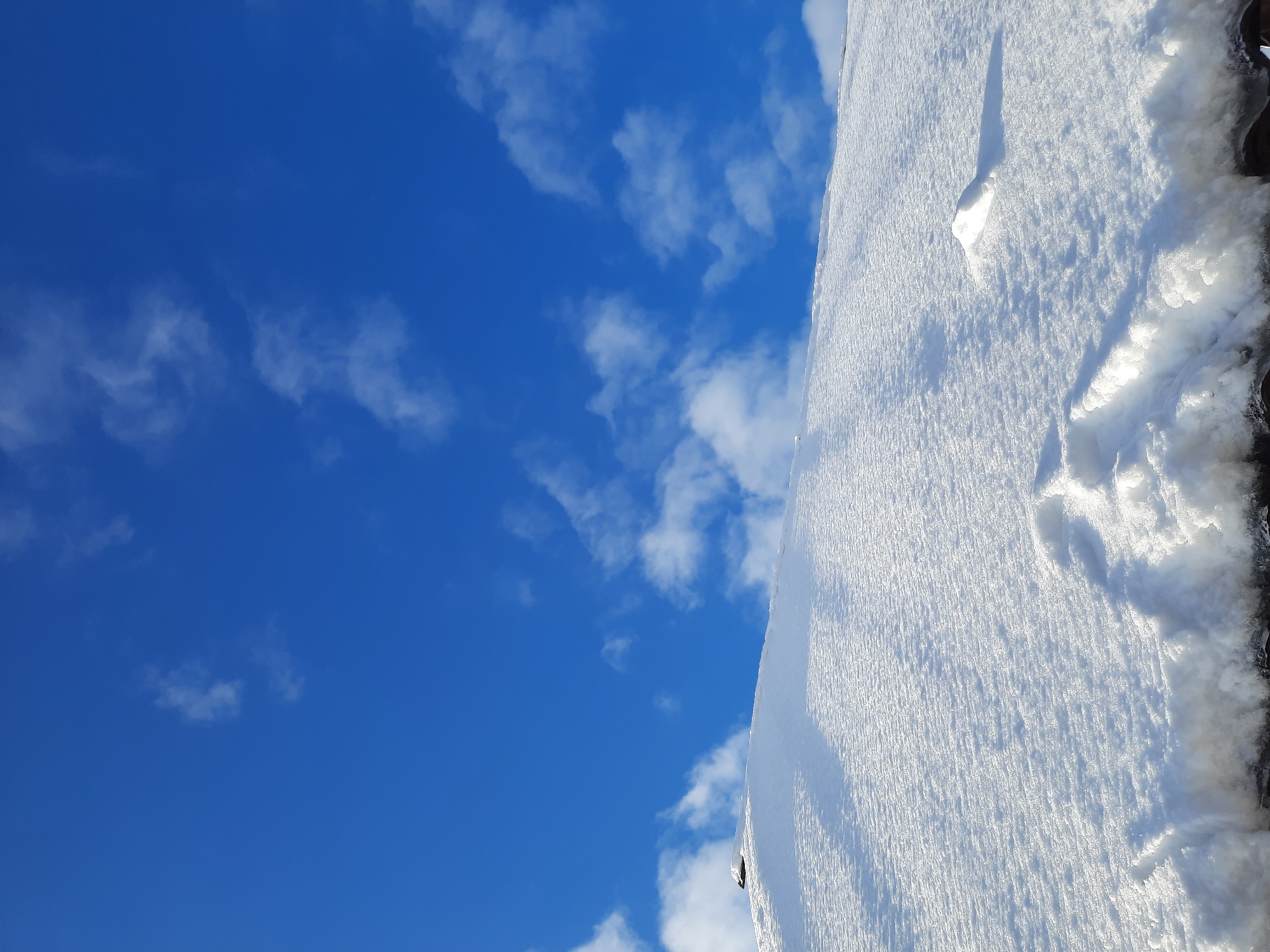 The image presents a bright and serene winter landscape under a clear blue sky, set in Krasnogorskoye, Russia. The scene is divided diagonally, with the upper-left portion dominated by a vibrant, deep blue sky adorned with scattered, wispy white clouds.
The lower-right section of the frame features an expansive surface covered in pristine, sparkling white snow. This snow-covered area, likely a rooftop or ground viewed from an upward angle, exhibits a distinct texture of fine ridges and glittering ice crystals, indicating direct sunlight. Two prominent indentations, possibly footprints, are visible in the snow. One is an elongated, distinct mark, casting a clear shadow that suggests a low sun angle. A smaller, more rounded impression is located further along the snowy expanse. The edge of the snow-covered surface, running along the bottom and right, shows some darker, rougher textures, possibly part of an underlying structure or iced-over sections.
The overall impression is one of a crisp, sunny winter day. There are no people visible, and no specific activity is taking place, beyond the evidence of a past presence in the footprints. No visible text is present in the image. The bright light and clear shadows indicate it is daytime.