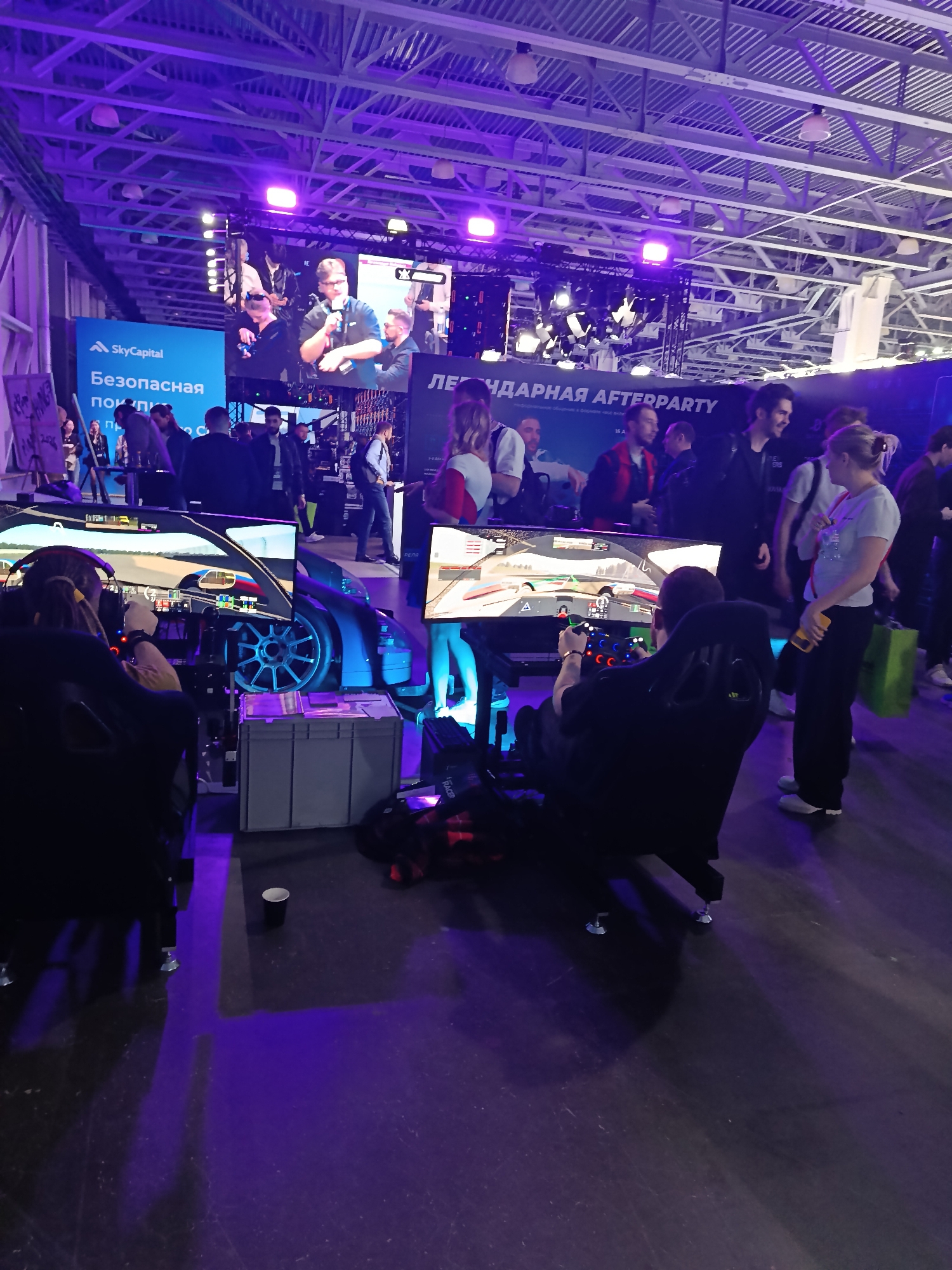 The image captures a scene at an event in an indoor location, possibly a convention center, illuminated with purple and blue lighting. There are several people present, including two individuals engaged in playing racing simulators. They are seated in gaming chairs in front of large monitors that display racing game visuals.
Around the simulators, there are people observing and interacting. In the background, a large screen displays content, possibly presentations or entertainment, with additional people gathered to watch.
Signage with text in Cyrillic can be seen in the background, along with other displays.
The location appears to be a public event, likely a gaming or technology convention.