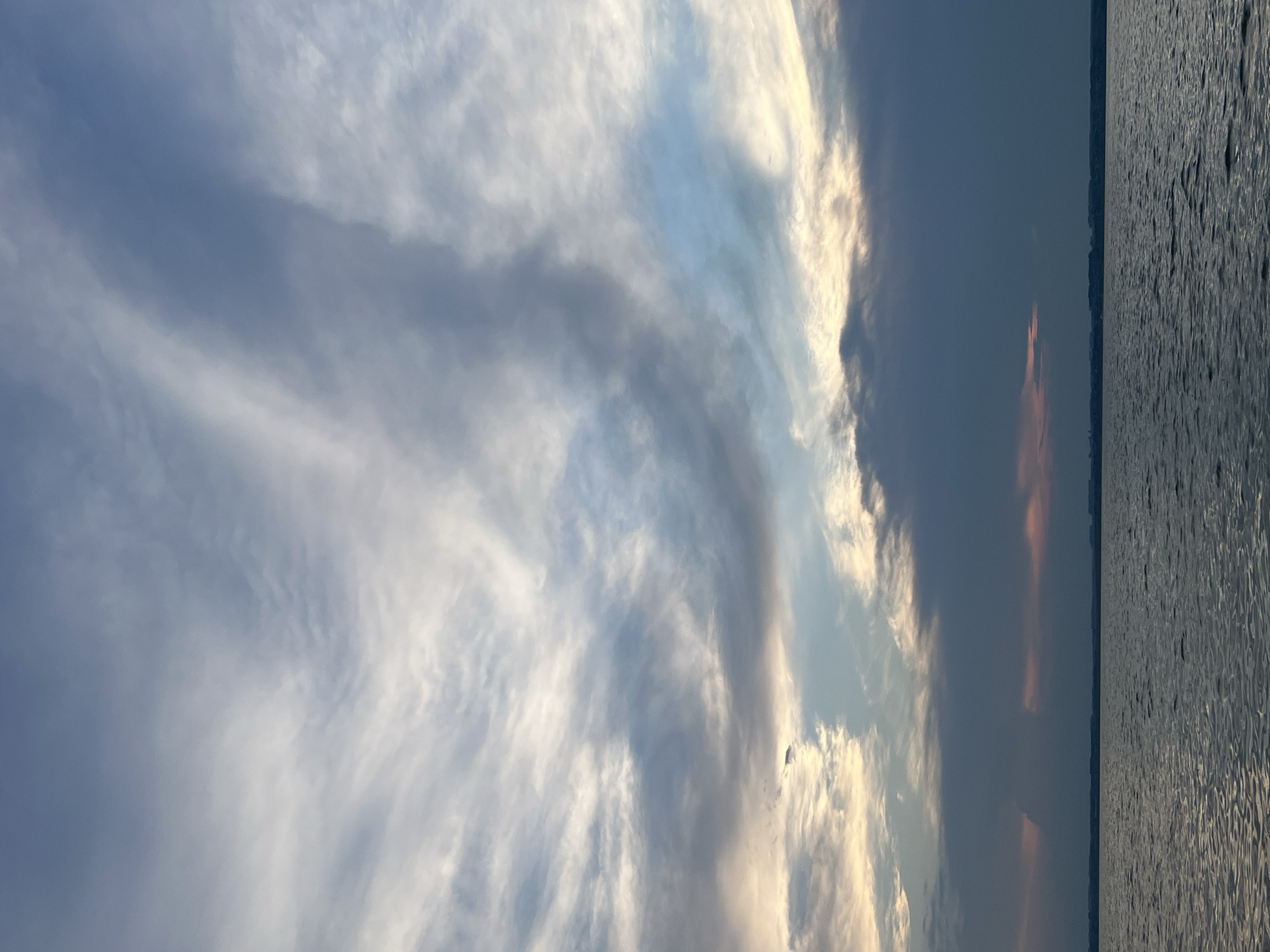 This is a vertical, low-angle, full shot of a body of water with a dramatic sky above. The water occupies the bottom third of the image and is dark and choppy, with ripples catching the light. The horizon line is visible, separating the water from the sky, and a faint, dark landmass can be seen in the distance. The sky is filled with a variety of clouds. The upper portion of the sky is a clear, pale blue, transitioning into wispy, white cirrus clouds that stretch across the frame. Below these, the clouds become thicker and darker, with shades of grey, blue, and hints of pink and orange near the horizon. The overall impression is of a moody and atmospheric sunset or sunrise. The location is identified as Novopichugovo, Russia. There are no people or specific objects visible in the scene.