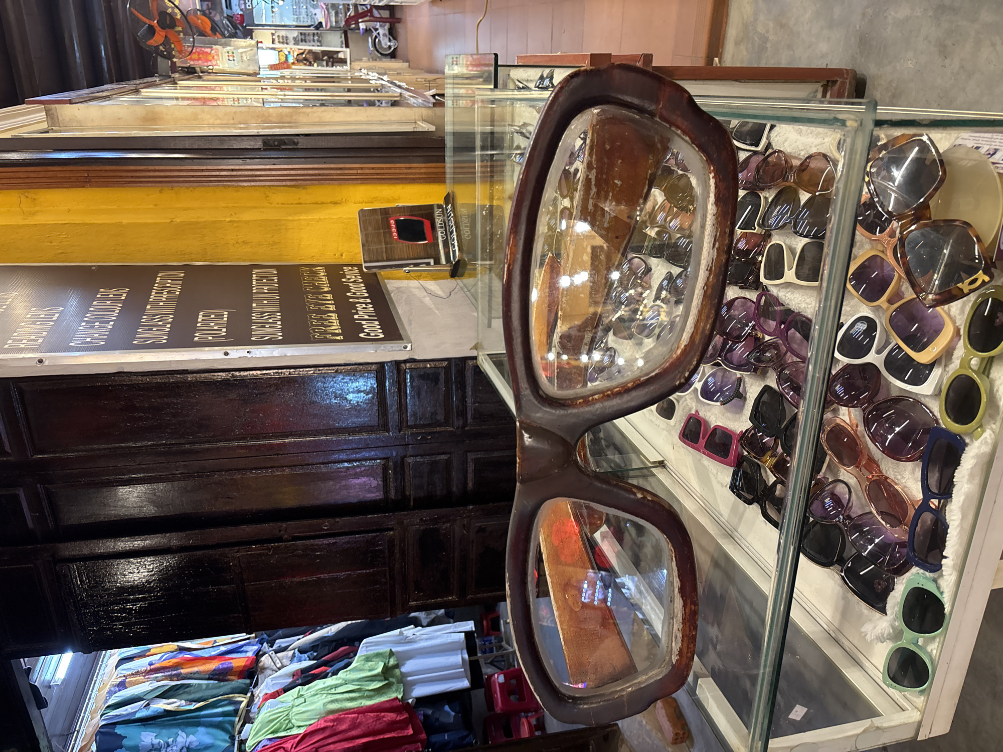 The image depicts a store display in Hội An Ward, Vietnam. A large, oversized pair of eyeglasses with brown frames dominates the foreground. The lenses are clear, reflecting the surroundings. Behind the large glasses is a glass display case containing various pairs of sunglasses arranged on a white surface. Behind this display case is a dark brown wooden cabinet with rectangular panels. A sign in front of the cabinet advertises sunglasses with prescription lenses (polarized). The sign also advertises a free eye check and promotes good prices and good service. Below the cabinet, folded clothing in various colors (green, red, blue) is visible. Further in the background, more shop displays and other objects are visible.