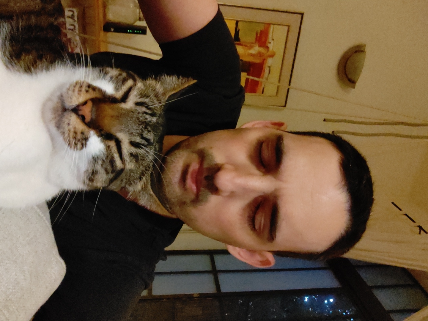 A horizontal, close-up shot depicts a man and a cat resting close together. The man, positioned to the right, has his eyes closed. He has dark hair, fair skin, and a stubbled beard. He wears a black, short-sleeved shirt. To his left, the cat also has its eyes closed. It is a mix of white and tabby (brown and black stripes) fur. The cat's face rests close to the man's neck. The background reveals a hint of an indoor setting. A picture hangs on the wall and a window offers a view of city lights at night, suggesting it may be in Tel-Aviv, Israel.