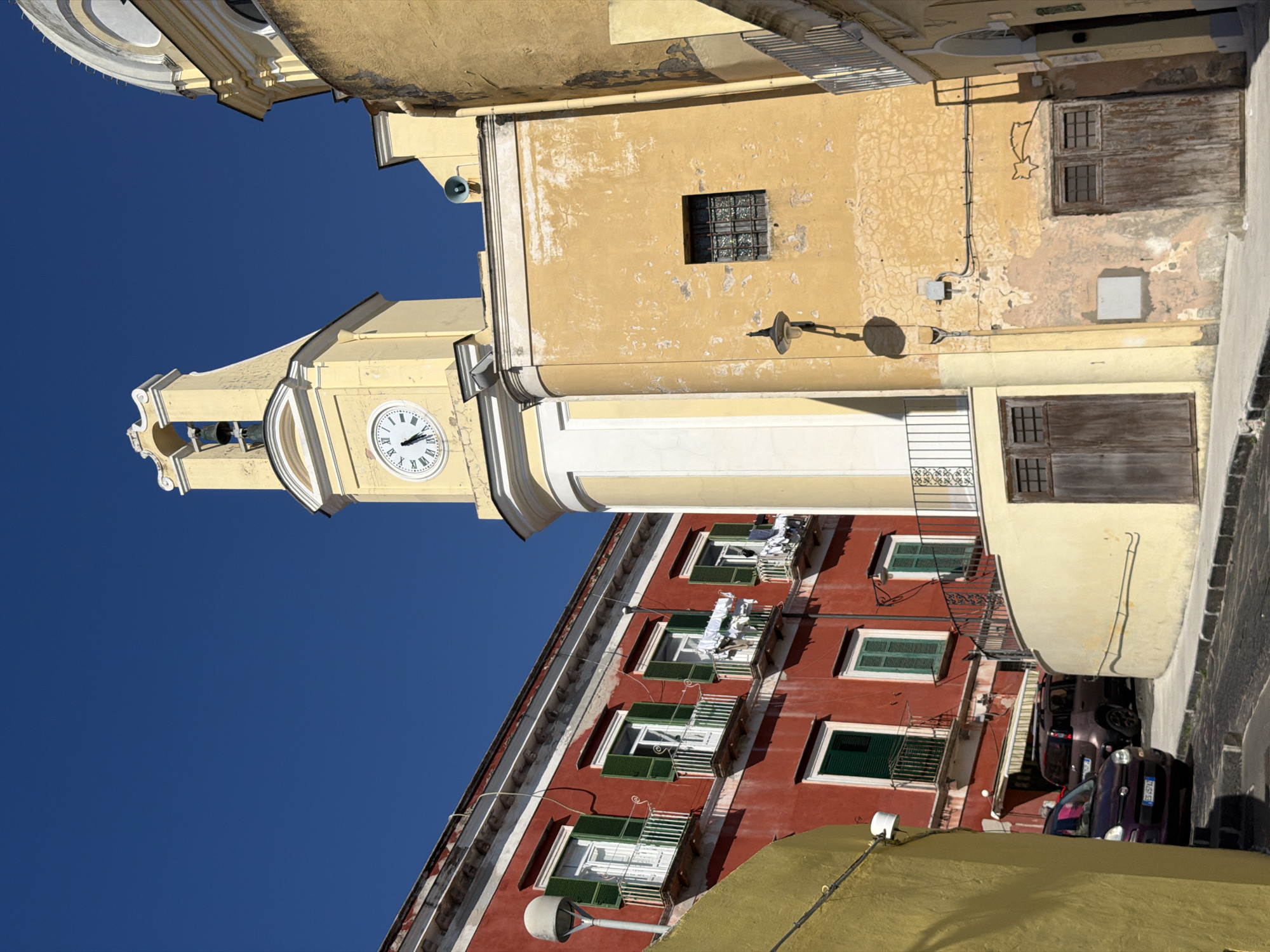 A low-angle shot, oriented at a slight angle, features a light yellow bell tower with a clock face against a clear blue sky. The clock face reads approximately 9:07. Adjoining the tower is a weathered yellow building with square windows, electrical conduits, and at least one exterior light fixture. Adjacent to this, a building with red exterior walls is noticeable. The windows on the red building are adorned with green shutters. Clothes hang on the balconies of the red building. On the side of the street, part of a sidewalk, a stone wall, and two parked cars are partially visible. The location of the image is Procida, Italy.