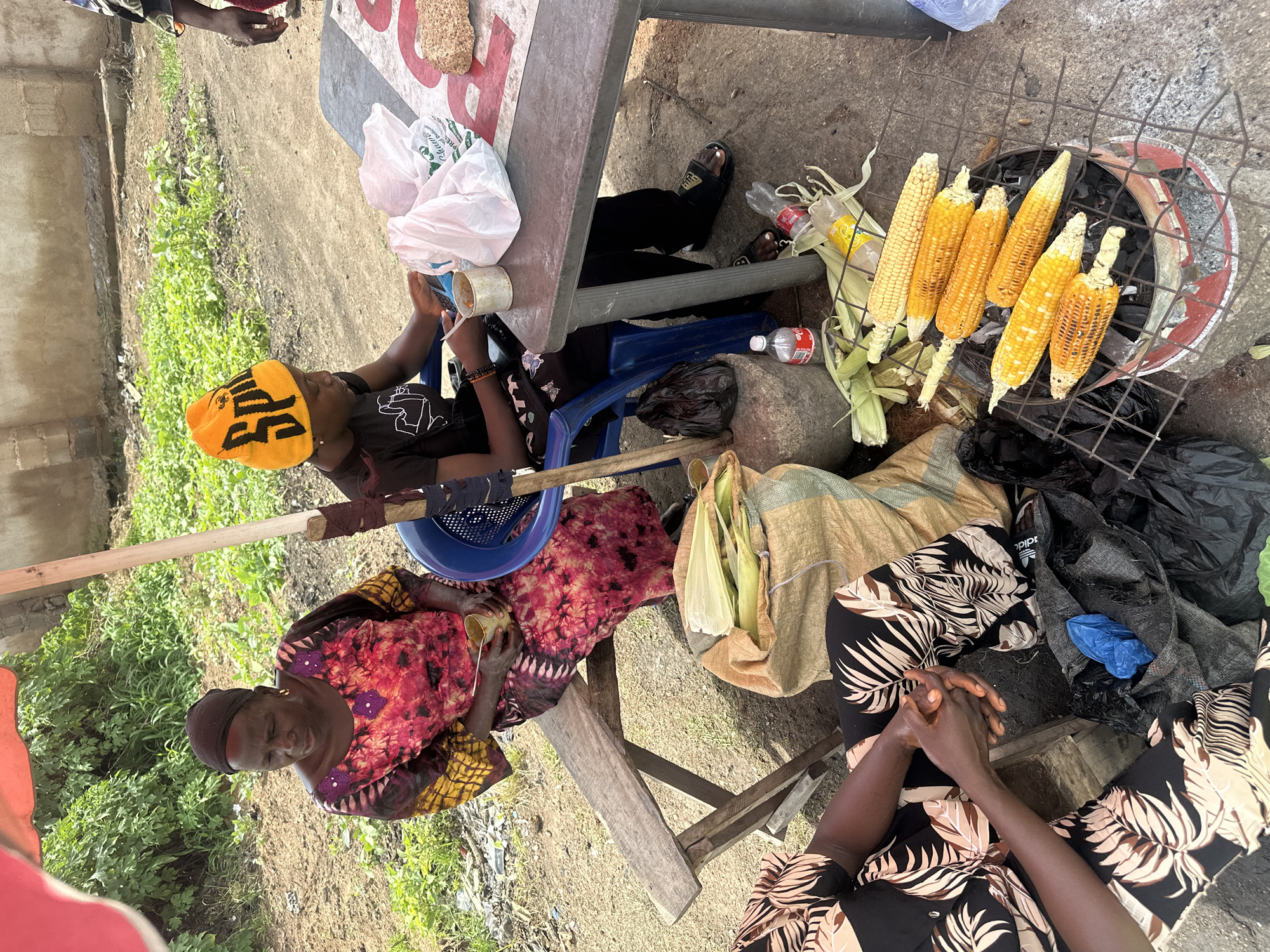 This media file depicts an outdoor scene in Gura Top, Nigeria, likely during daylight hours given the bright illumination. The primary subjects are three individuals, two women and a young person, engaged in a transaction or preparation of food.
In the foreground, a woman wearing a vibrant, multi-colored patterned garment is seated, looking down with her hands clasped. Beside her, a young person wearing a bright yellow beanie with black lettering sits on a blue plastic chair, holding a small cup. Further back, another woman, mostly obscured, is also seated.
The central object of activity appears to be a grill laden with corn on the cob, actively cooking over charcoal. Several unhusked corn cobs are also visible, suggesting they are part of the preparation or sale. Scattered around the scene are various objects including plastic bottles, bags, and a blue plastic basin.
The setting is an outdoor area with a rough ground surface and some sparse vegetation. A table with a patterned top is present, as well as wooden crates and what appears to be a makeshift stall structure. The overall atmosphere seems to be that of a street food vendor.
There is visible text on the yellow beanie worn by the young person, though it is not entirely legible. The scene suggests a casual, everyday interaction centered around the sale and preparation of corn.