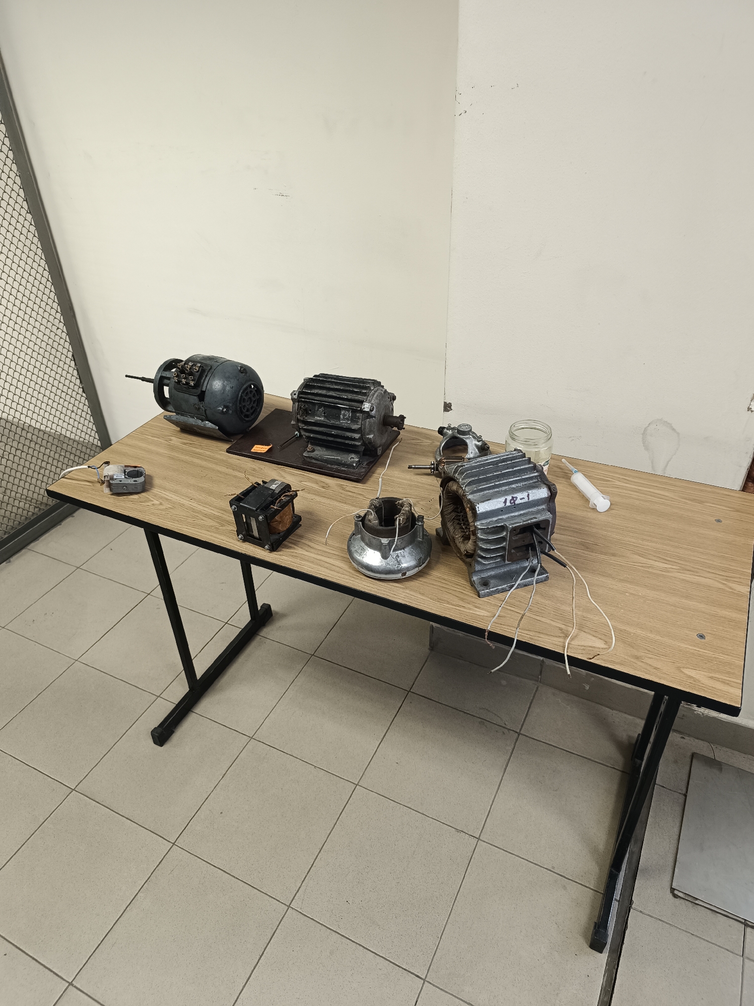 The image shows a room with tiled floors and light-colored walls. A black metal cage is visible in the left background. A wooden table with black legs stands in the center. On the table are several disassembled electrical components, including three motors of different sizes, a transformer, a small electronic device, a glass jar, and a syringe. "18-1" is printed on one of the motor components. No people are visible. The setting suggests a workshop or laboratory environment. The location in Sofia, Bulgaria cannot be determined from the image.