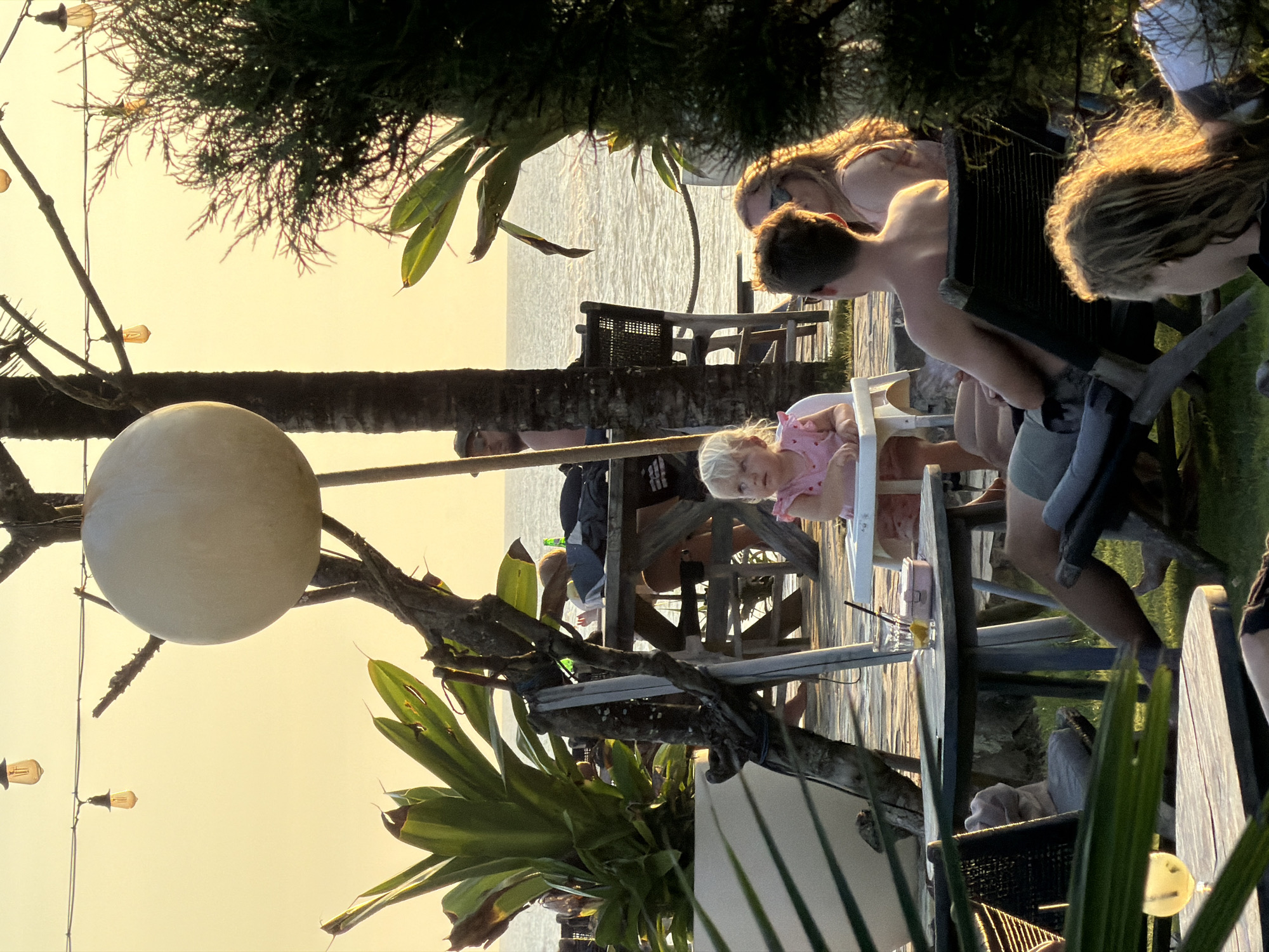 This image captures a family gathering at sunset, likely in Mengwi, Indonesia, given the tropical flora. A young girl in a pink dress sits in a white highchair, seemingly observed by a boy standing next to her and a woman. Other adults are seated around a table. Overhead, a large, spherical light fixture hangs from a tree, and string lights add to the ambiance. The scene overlooks water, possibly the ocean or a large lake, with other tables and chairs visible in the background. The overall atmosphere is relaxed and summery.