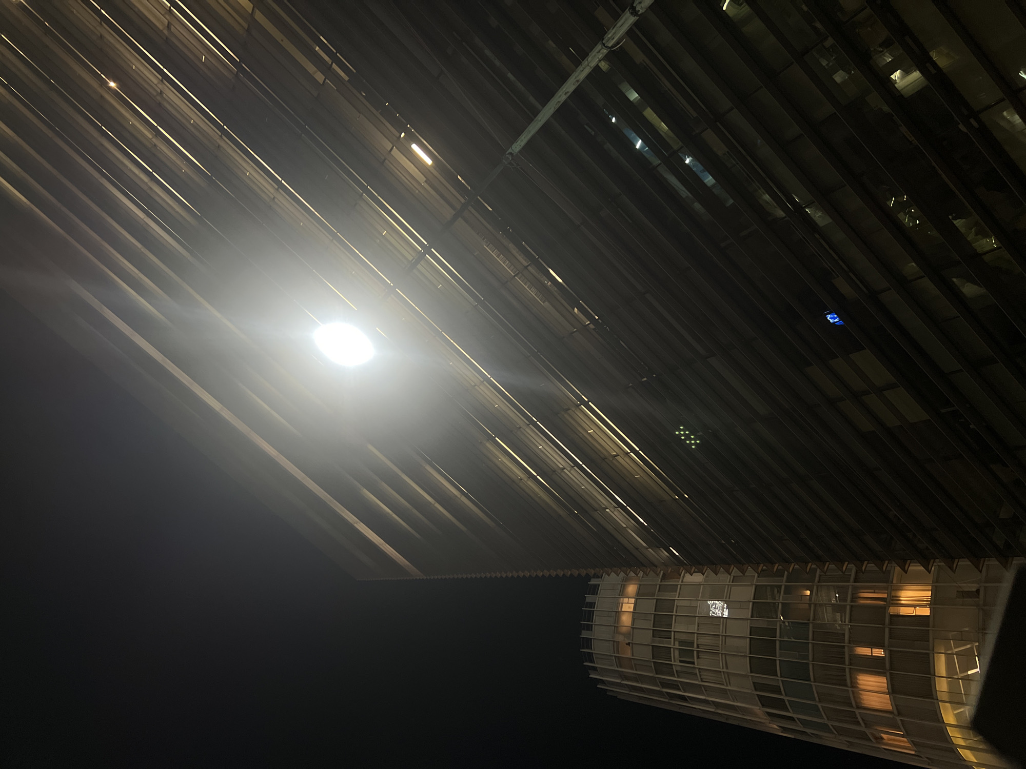 A low-angle night shot captures a modern architectural structure. The building's facade features a pattern of parallel, dark-toned slats or beams extending diagonally across the frame, with light visible through gaps. A bright spherical light source creates lens flare and obscures some details. Below the slats, a portion of a rounded structure with a grid-like pattern of windows is visible. Some windows are illuminated from within, casting a warm glow. The background is dark, suggesting a night scene in an urban environment. Considering the architectural features, the image seems to be of Soumaya Museum located in Mexico City, Mexico.