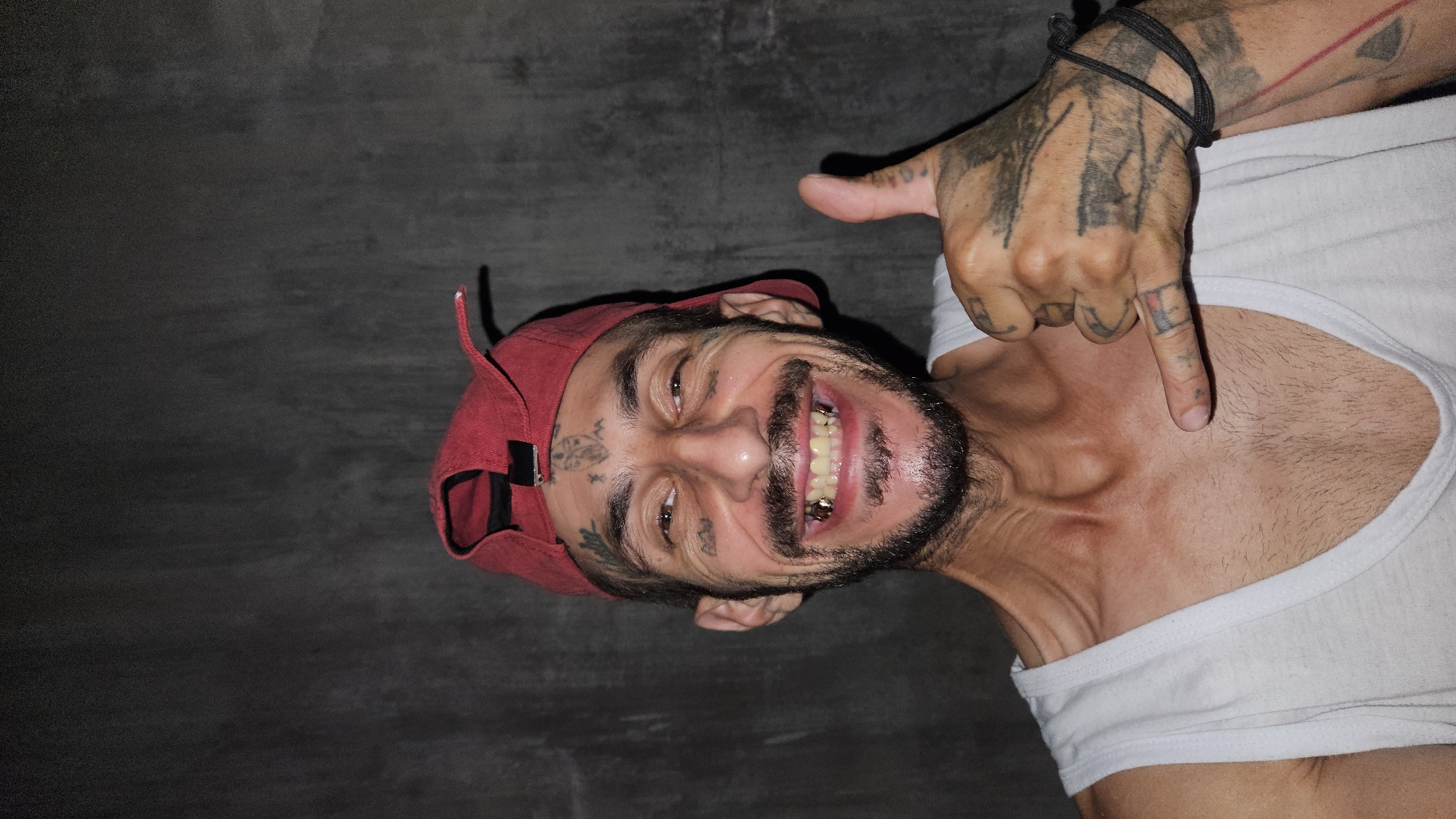A man with a cheerful, wide smile, showcasing gold caps on his lower teeth, is captured in a close-up, rotated portrait in Cam Đức, Vietnam. He is wearing a white tank top and a red baseball cap, worn backward. His head is tilted, and the image is rotated approximately 90 degrees clockwise, making his body appear horizontal against a dark, featureless background, possibly a wall or floor.
The man has a full dark beard and mustache, along with extensive tattoos on his face, including around his eyes and forehead, as well as on his hands and arms. His right hand, adorned with more tattoos, is raised with the thumb pointing upwards and the index finger pointing downwards towards his upper chest/collarbone area. His expression is one of joy or amusement, with his eyes sparkling. The dim lighting of the scene suggests it might be nighttime or an indoor setting. No discernible text is visible on his clothing or in the background, though some tattoos might contain illegible script or symbols.
