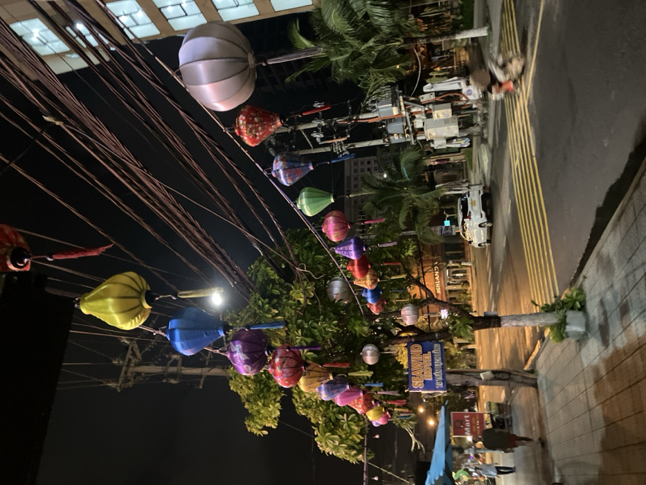 The image depicts a nighttime street scene in Ngũ Hành Sơn Ward, Vietnam. The foreground features a concrete sidewalk with two pedestrians; one individual in a dark shirt and red shorts walks away from the camera, while another in a lighter shirt is partially visible further down. To the left, a blue awning covers an outdoor stall, and a red "Mart" sign is visible. A tree central to the sidewalk is heavily decorated with numerous multicolored silk lanterns of various shapes, including yellow, blue, purple, red, green, and silver. A blue sign for "SEAWIND HOTEL, 10 TRẦN BẠCH ĐẰNG" is attached to the tree.
The midground shows a paved two-lane road with distinct yellow horizontal striped markings. A white passenger vehicle is parked on the right side of the road, and a motion-blurred scooter with a rider is traversing the road from right to left. Overhead, a dense network of black electrical wires crisscrosses the dark night sky, with additional lanterns strung between poles and across the street. A street light is also visible. In the background, a multi-story building with illuminated windows stands on the right, and palm trees are interspersed along the street and near the buildings. A "HIGHLANDS COFFEE" sign is visible in the distance.