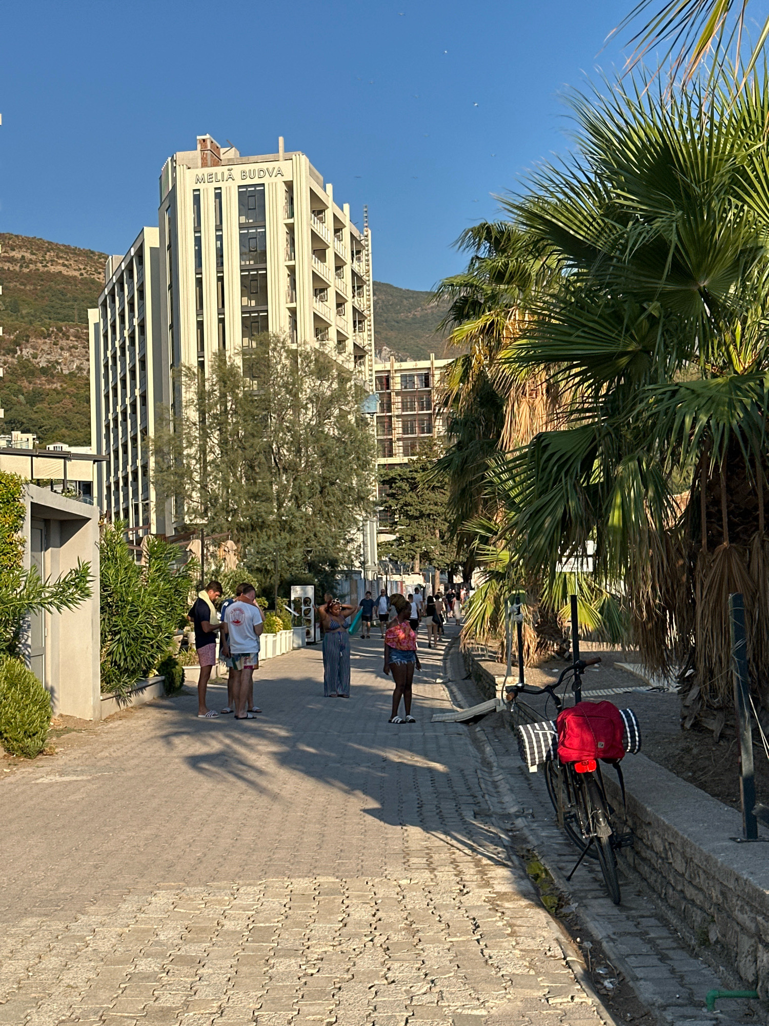 A street scene in Rafailovići, Montenegro, features the Melia Budva hotel on the left, a tall, cream-colored building with many windows and balconies, set against a backdrop of a treed, rocky hillside. In the foreground, a paved walkway is lined with low greenery on the left and palm trees on the right. Several people are walking on the walkway. A bicycle with a red bag and a rolled-up striped item is parked on the right side of the walkway. The sky is clear and blue.