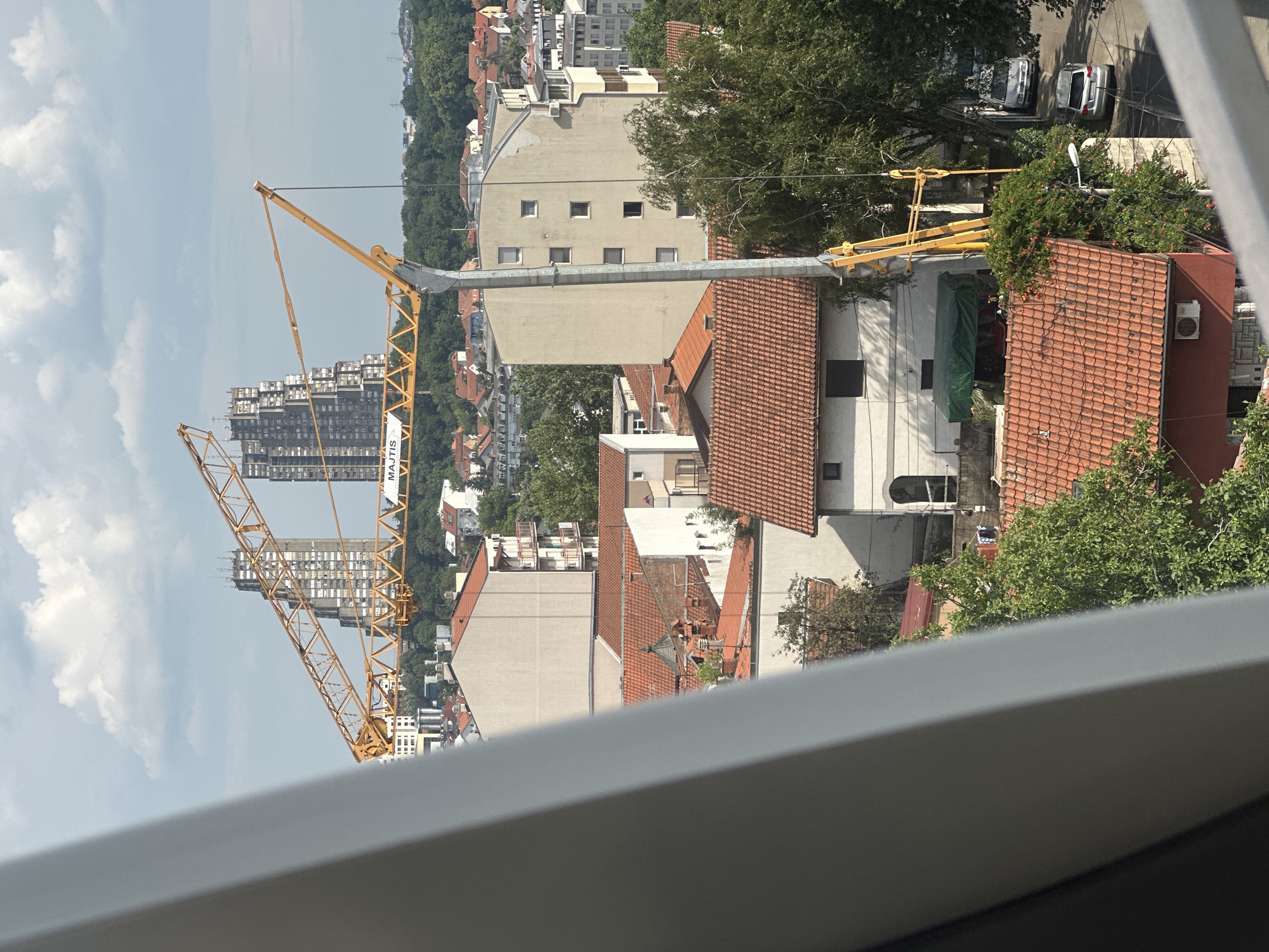 This is a vertical, high-angle, full shot of an urban construction scene in Belgrade, Serbia, under a bright, partly cloudy sky, suggesting midday.
The primary focus is a large yellow construction crane dominating the left and center of the frame. Its latticed structure extends diagonally upwards. In the background, a tall, grey, modern apartment building with numerous windows is under construction, partially obscured by the crane. Further in the distance, more buildings and greenery are visible, indicating a densely populated urban area with interspersed trees.
In the foreground and mid-ground, several residential buildings with red-tiled roofs are present. One building in the mid-ground features a beige facade with multiple small, square windows. Another building, to the right, has a red brick facade. The presence of construction materials, including a green tarp covering something on a rooftop, suggests ongoing building work. In the upper right portion of the image, two cars are parked on a street lined with trees.
The scene conveys a sense of urban development and ongoing construction activity. There is no visible indication of people. The only legible text is "MAJTIS" printed vertically on a white banner attached to the crane's structure.