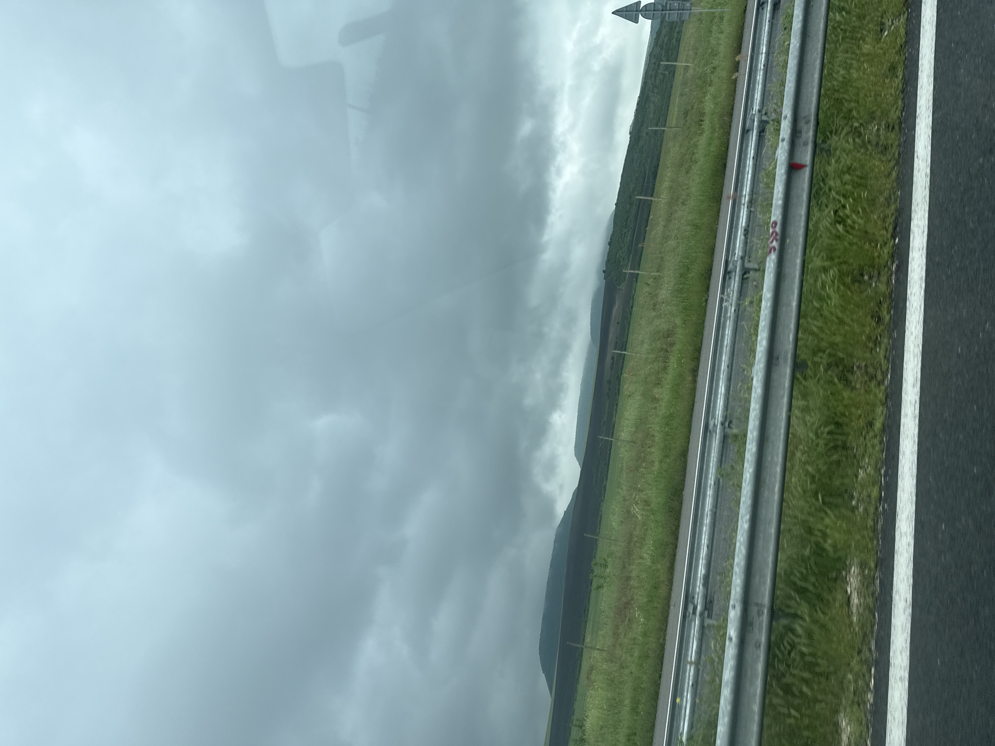 The image depicts a view from a vehicle traveling along a highway in Venchan, Bulgaria. The scene includes a roadway with a white painted line delineating the lanes. A silver-colored guardrail runs parallel to the road, separating the roadway from a grassy area. Beyond the grassy verge, there is a view of mountains under a heavily overcast sky. There is a sign visible in the upper part of the image. The scene suggests a rural or semi-rural setting.
